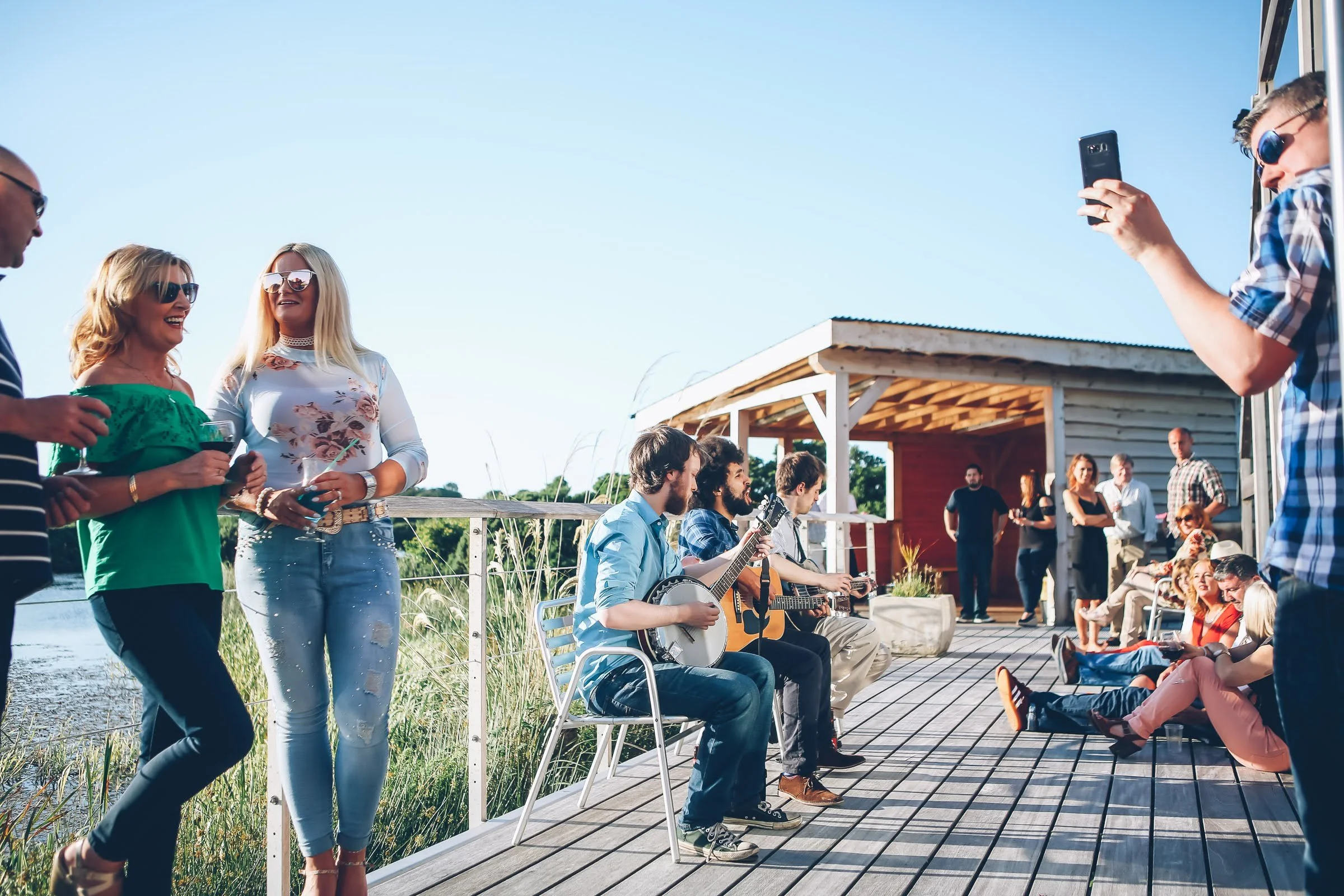People enjoying an outdoor gathering beside lake, with musicians playing guitars and others socialising, under a clear blue sky.