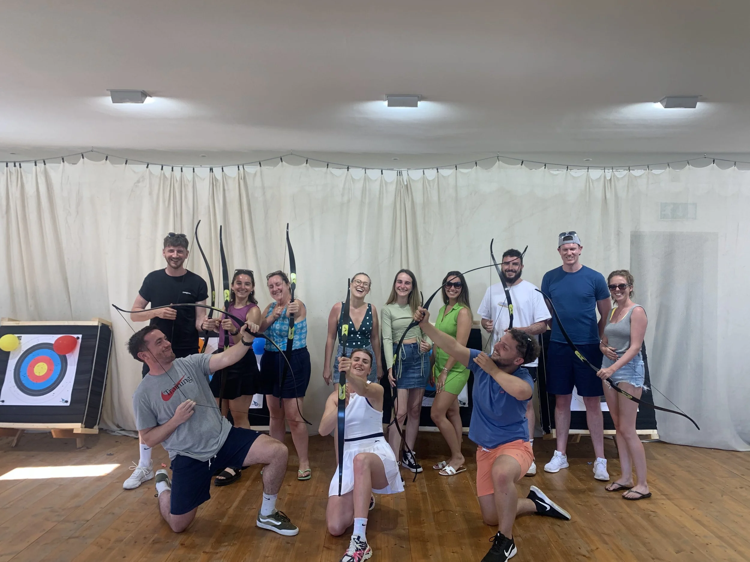 A group of people posing with archery bows and arrows indoors, standing in front of a beige curtain, with a target on the left side of the image, smiling and celebrating.