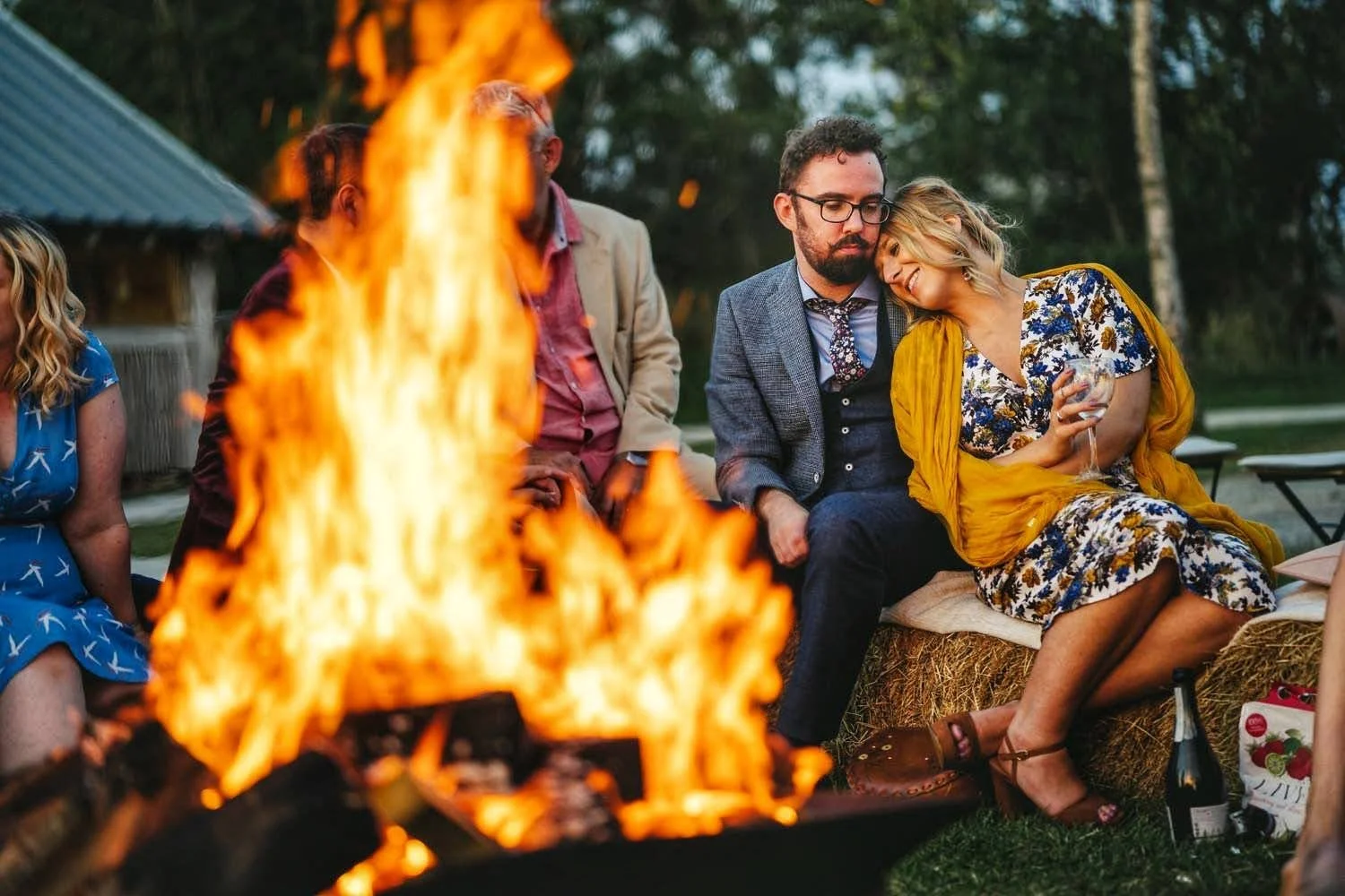 People sitting around a campfire at an outdoor gathering, with a woman resting her head on a man's shoulder, holding a wine glass, smiling, in a natural setting during the evening.