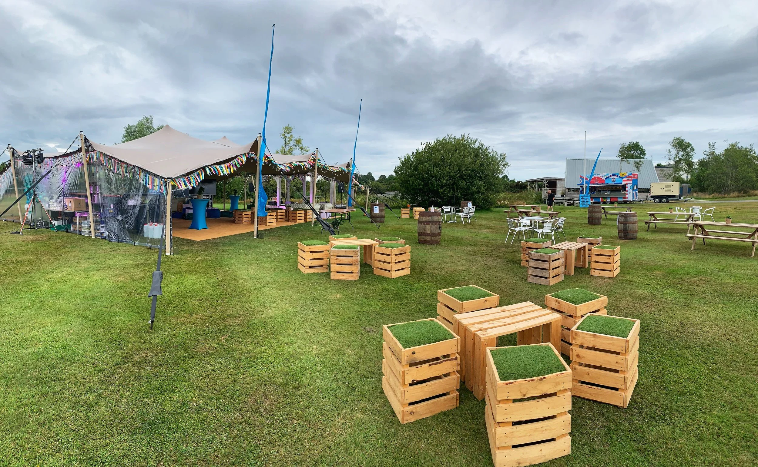 Outdoor event setup with large white tent decorated with colorful streamers, surrounded by wooden tables and benches, food truck in the background, and cloudy sky overhead.