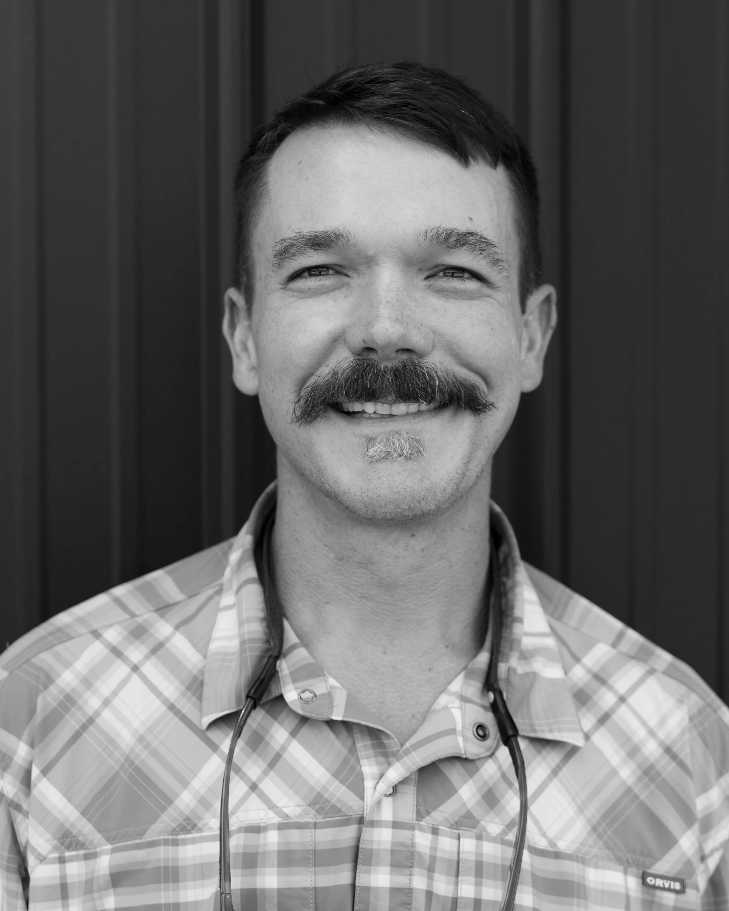 Black and white photo of a smiling man with short hair and a mustache, wearing a checkered shirt and a stethoscope around his neck, standing in front of a dark wooden wall.