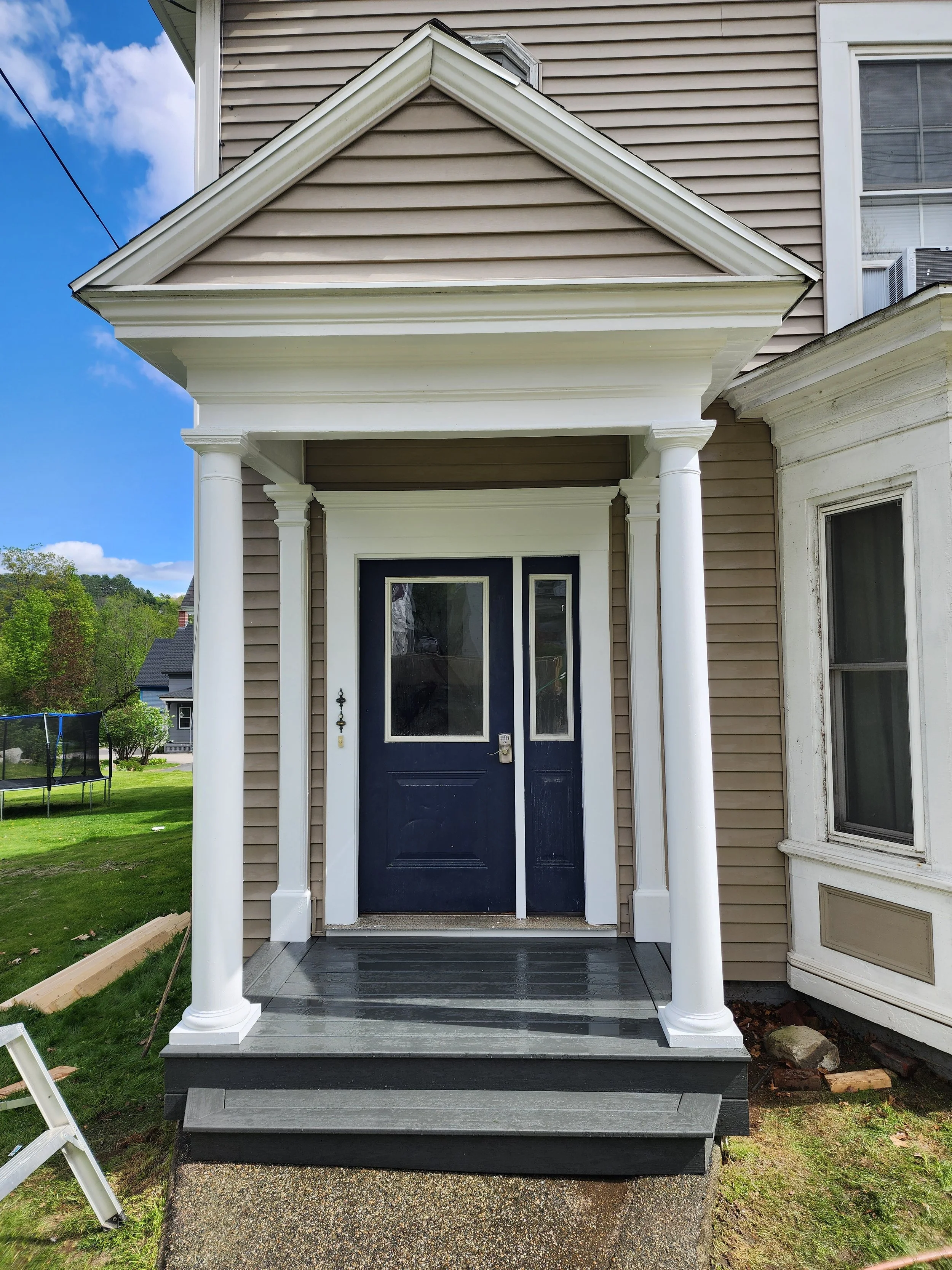 Front porch of a house with a black front door, white columns, gray steps, and vinyl siding, under a gabled roof with white trim, on a bright day.