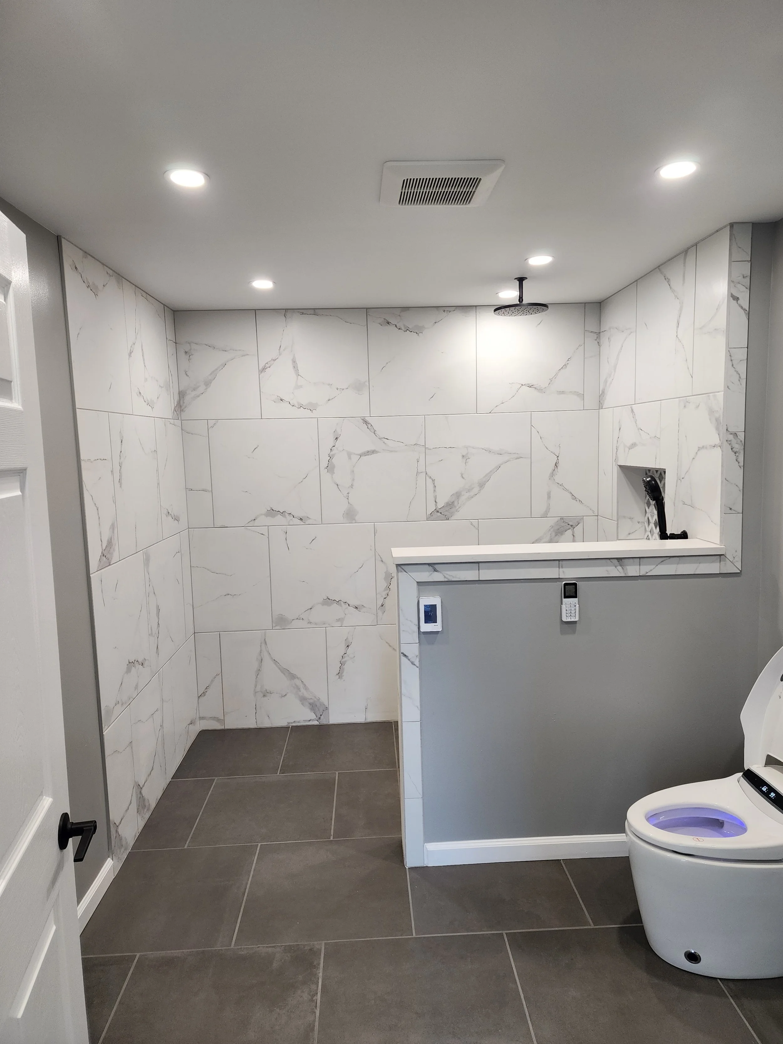 Modern bathroom with large marble tile walls, gray floor tiles, ceiling spotlights, a ceiling vent, and a high-tech toilet. There is also a small half-height wall with a built-in control panel and a surface for a sink.