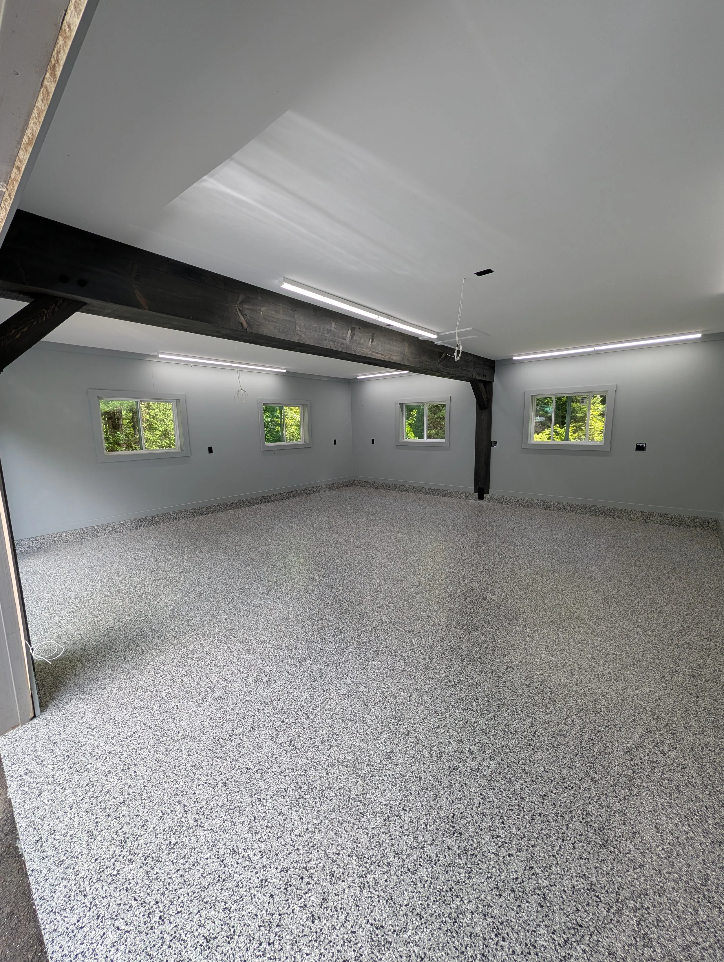 Interior of a room under renovation with a speckled gray carpet, white walls, multiple windows showing outdoor greenery, and exposed structural beams.