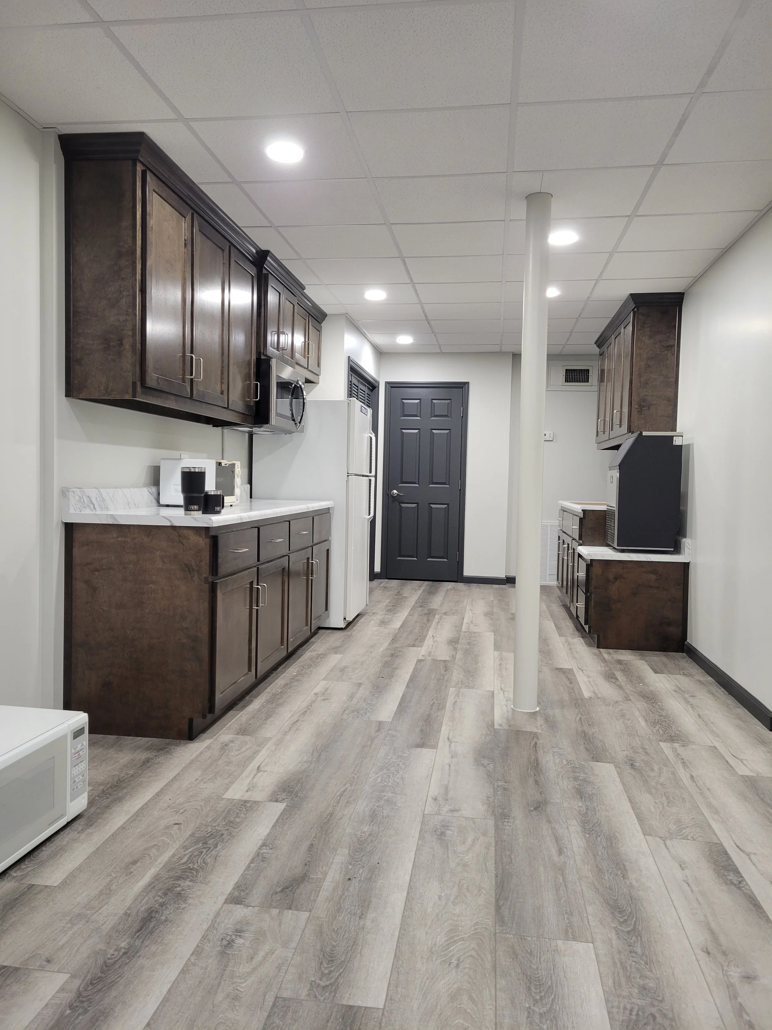 A kitchen with dark wooden cabinets, white countertops, a white refrigerator, microwave, and a small white appliance, with wood-look flooring, a dark door, and a column in the center of the room.