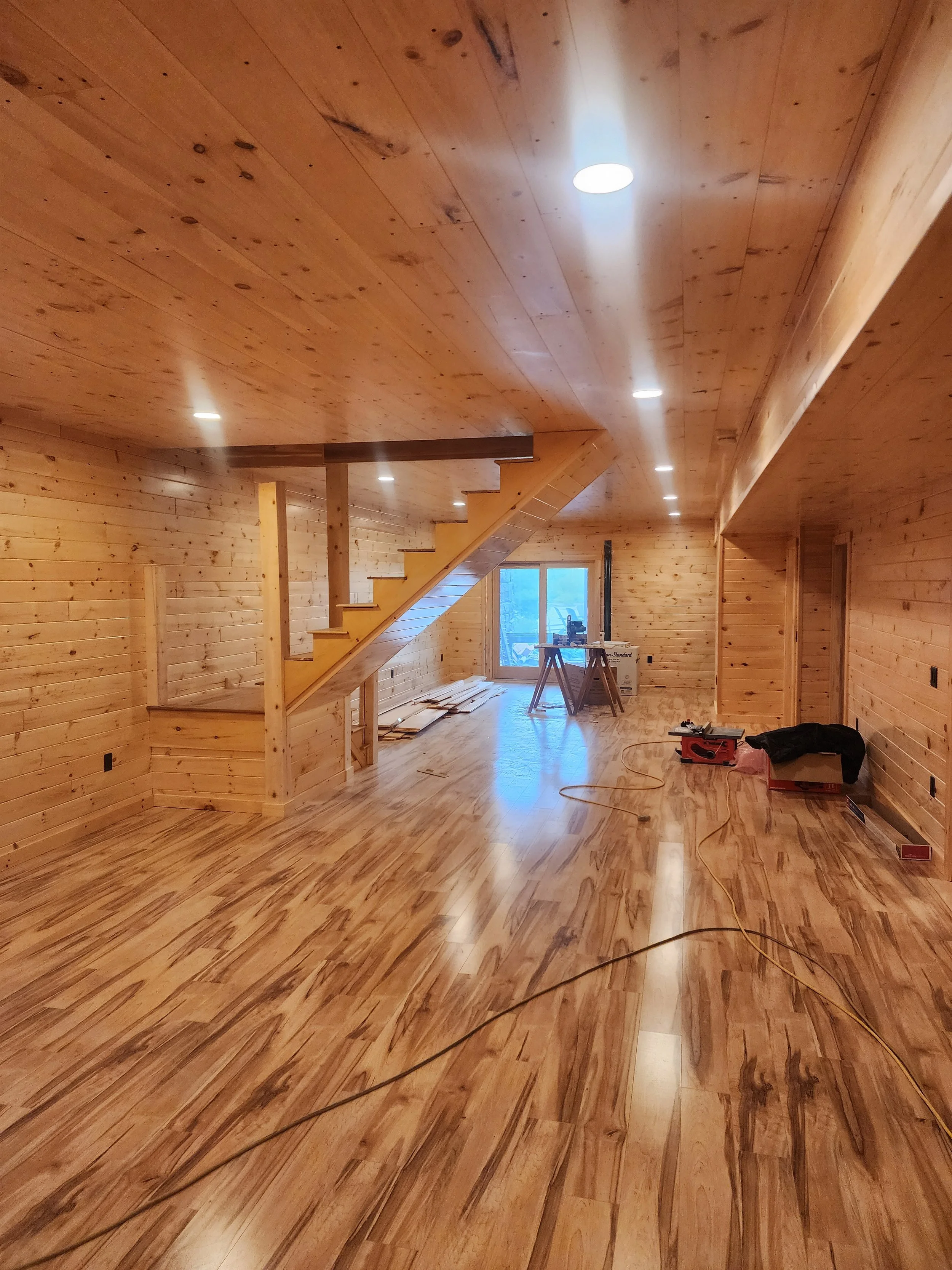 Interior view of a wooden house under construction. Finished wooden walls, ceiling, and floors, with construction tools and materials present, including a table saw, a box, and a ladder in the background.