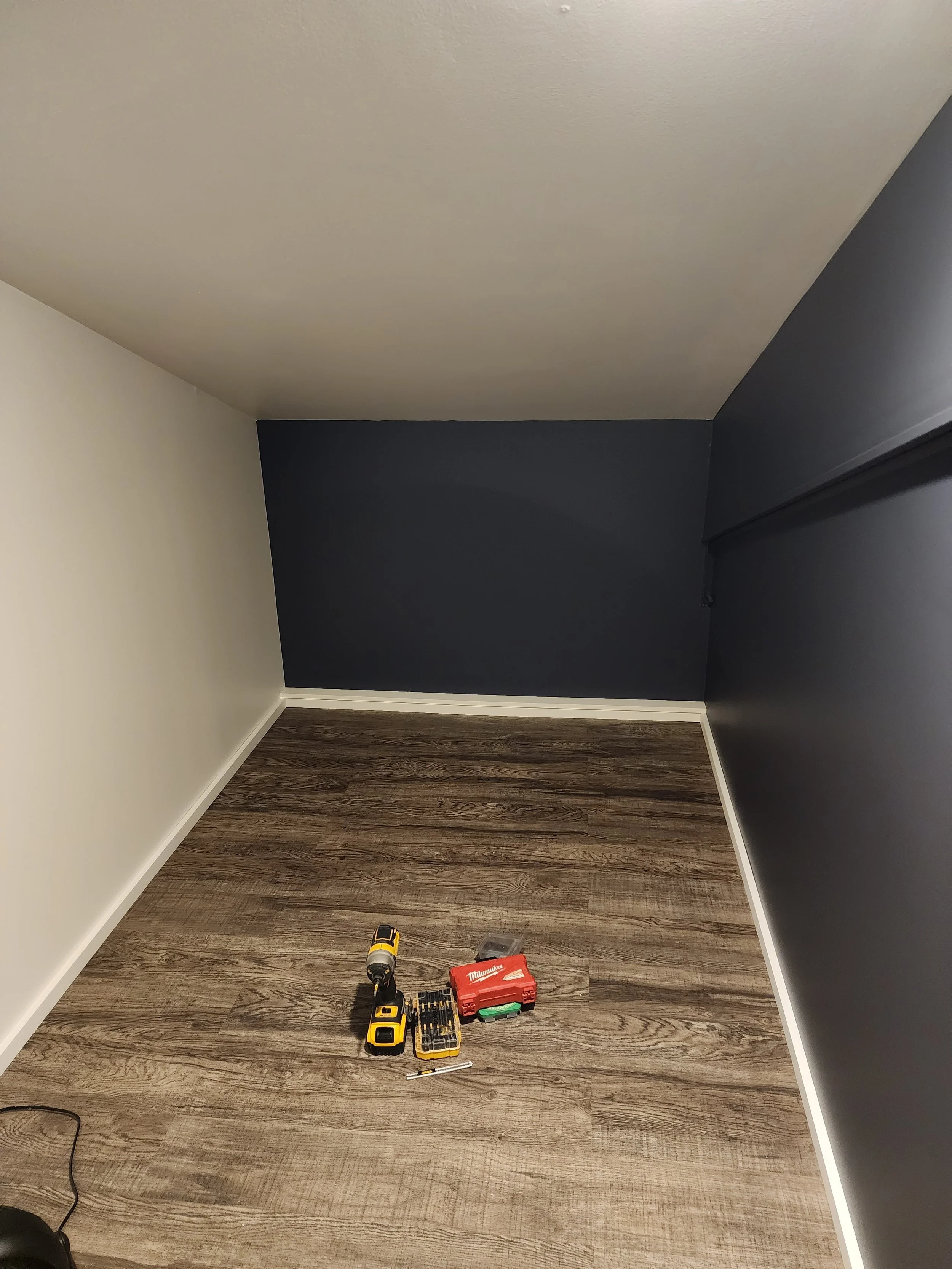 Empty corner of a room with dark blue and white walls, a hardwood floor, tools and a red toolbox on the floor, and a cordless drill.