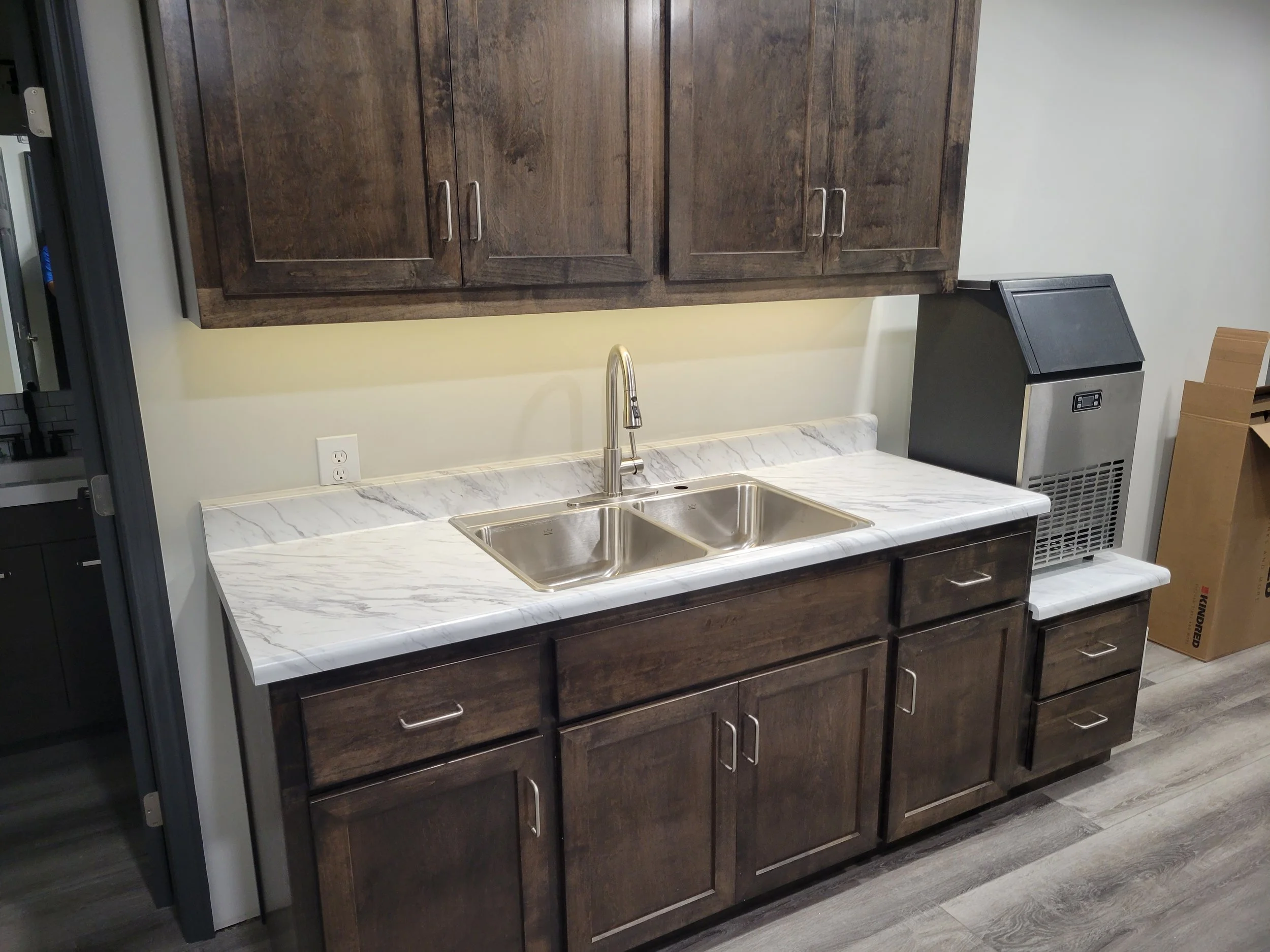 Kitchen with dark wood cabinets, a marble countertop, a stainless steel double sink, a paper towel holder, and a stainless steel appliance, possibly a small freezer or ice maker, to the right of the sink.