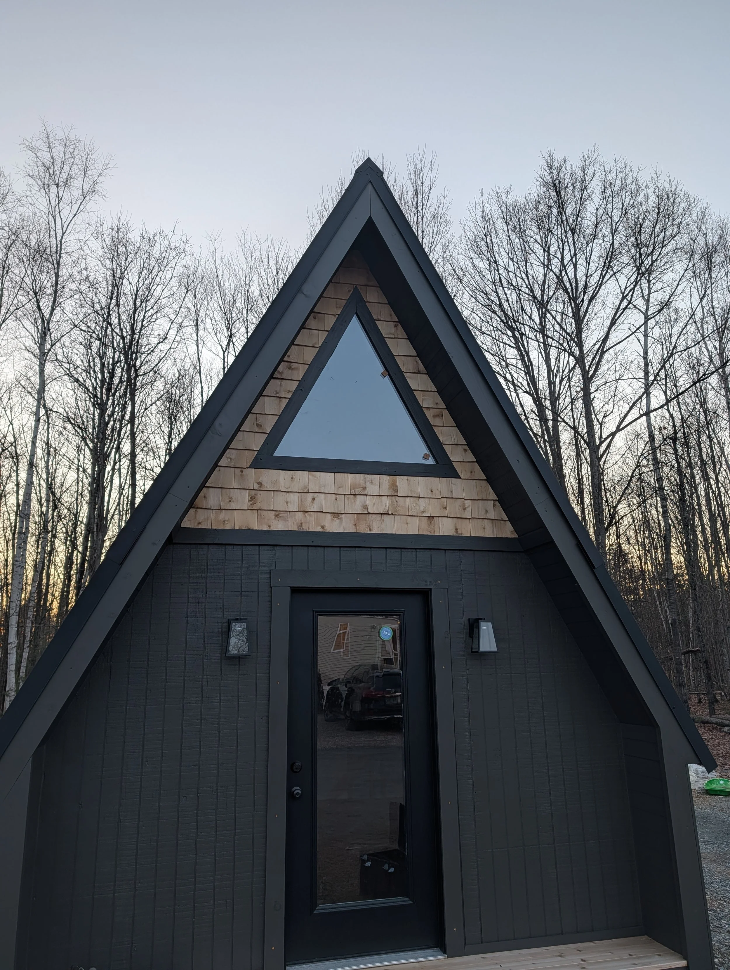 A small A-frame house with black exterior and wooden shingles on the upper section, featuring a triangular window and door, surrounded by leafless trees during sunset or sunrise.