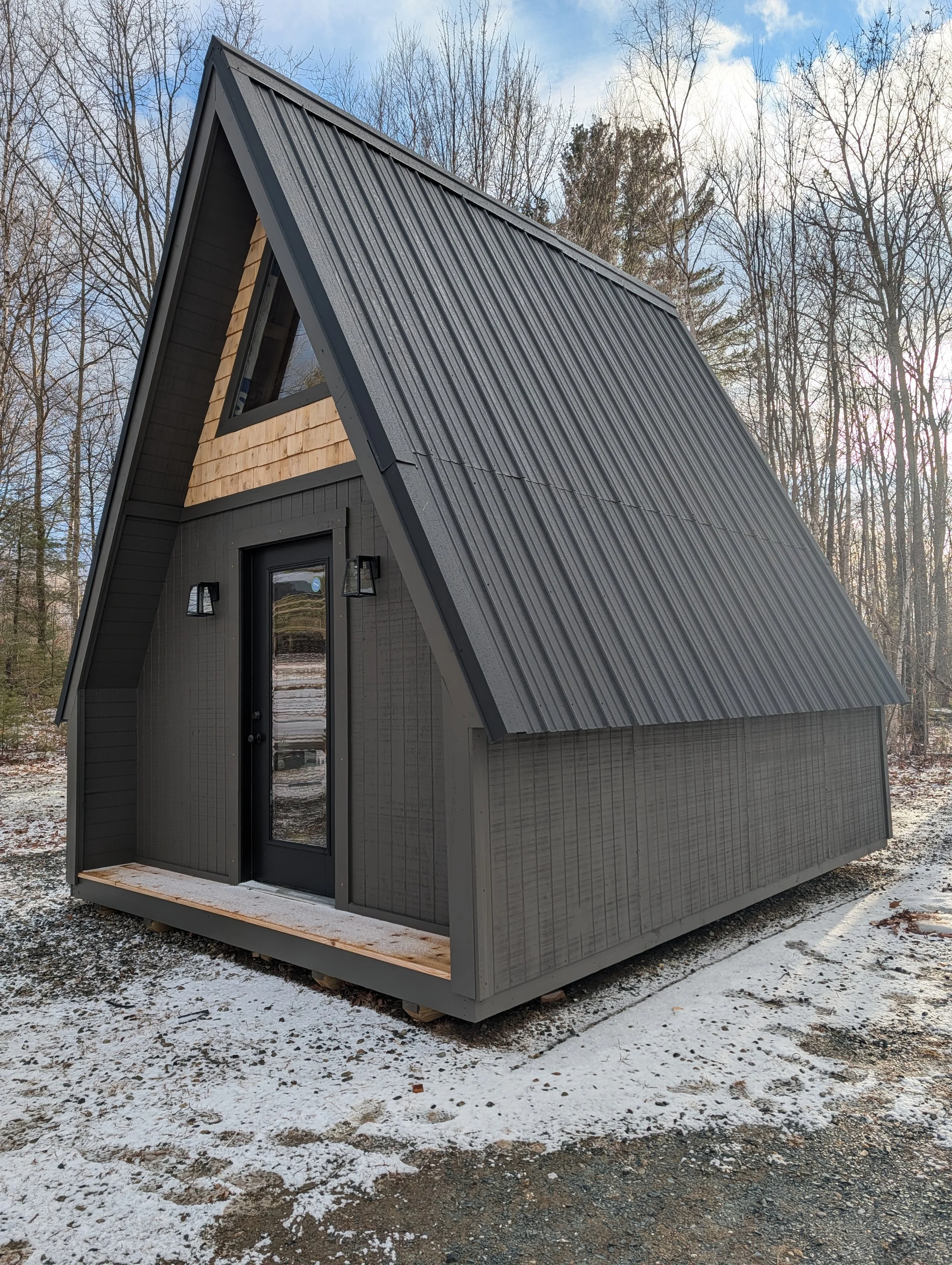 A tiny house with a curved metal roof, black exterior, and a small porch in a wooded area with snow on the ground.