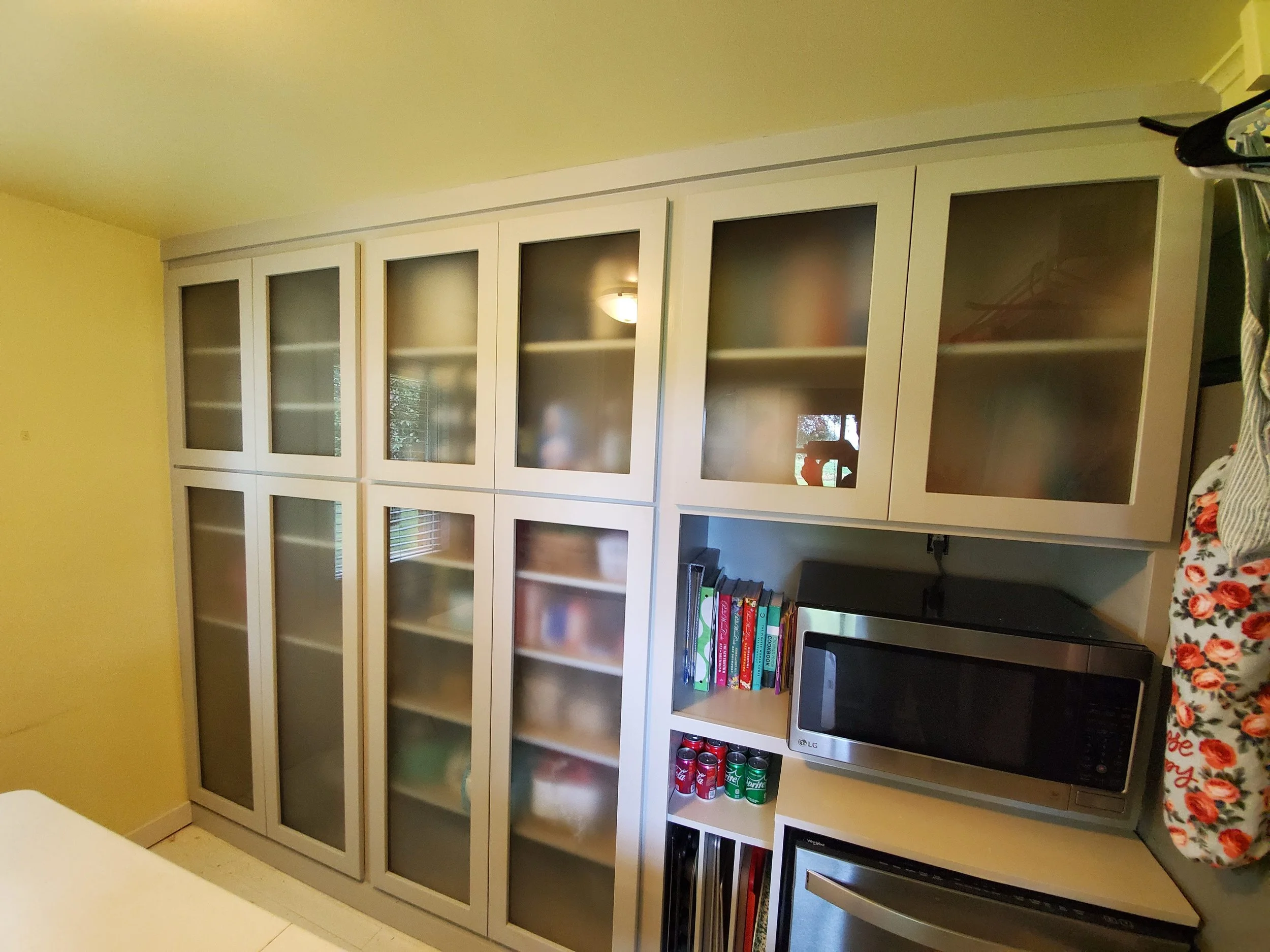 White cabinet with glass doors on upper shelves, filled with various items, in a kitchen with yellow walls. Below, a section holds books, a microwave, and soda cans.