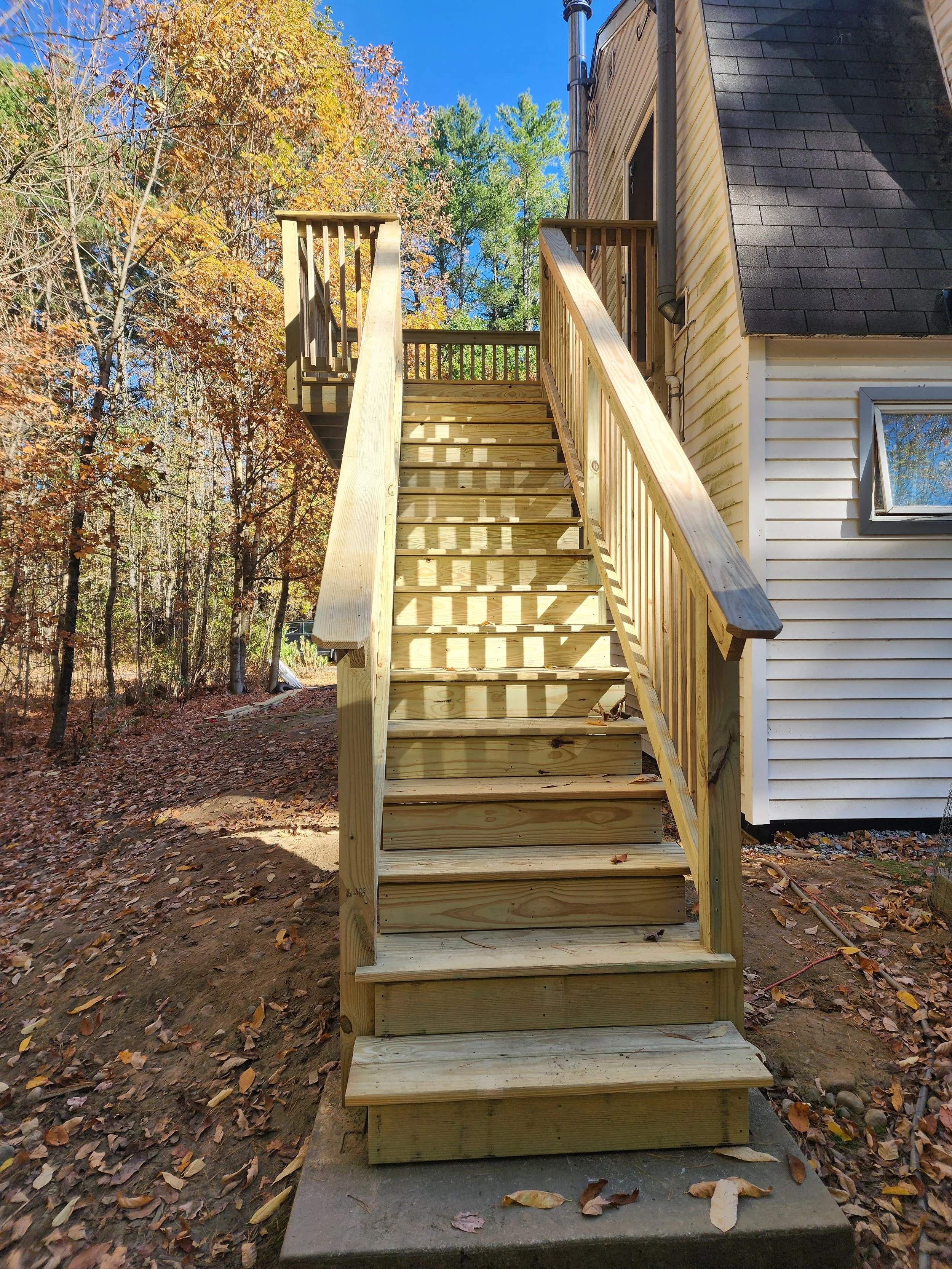 Wooden outdoor staircase leading to a house door, with trees and fallen leaves in the background during fall, under a clear blue sky.