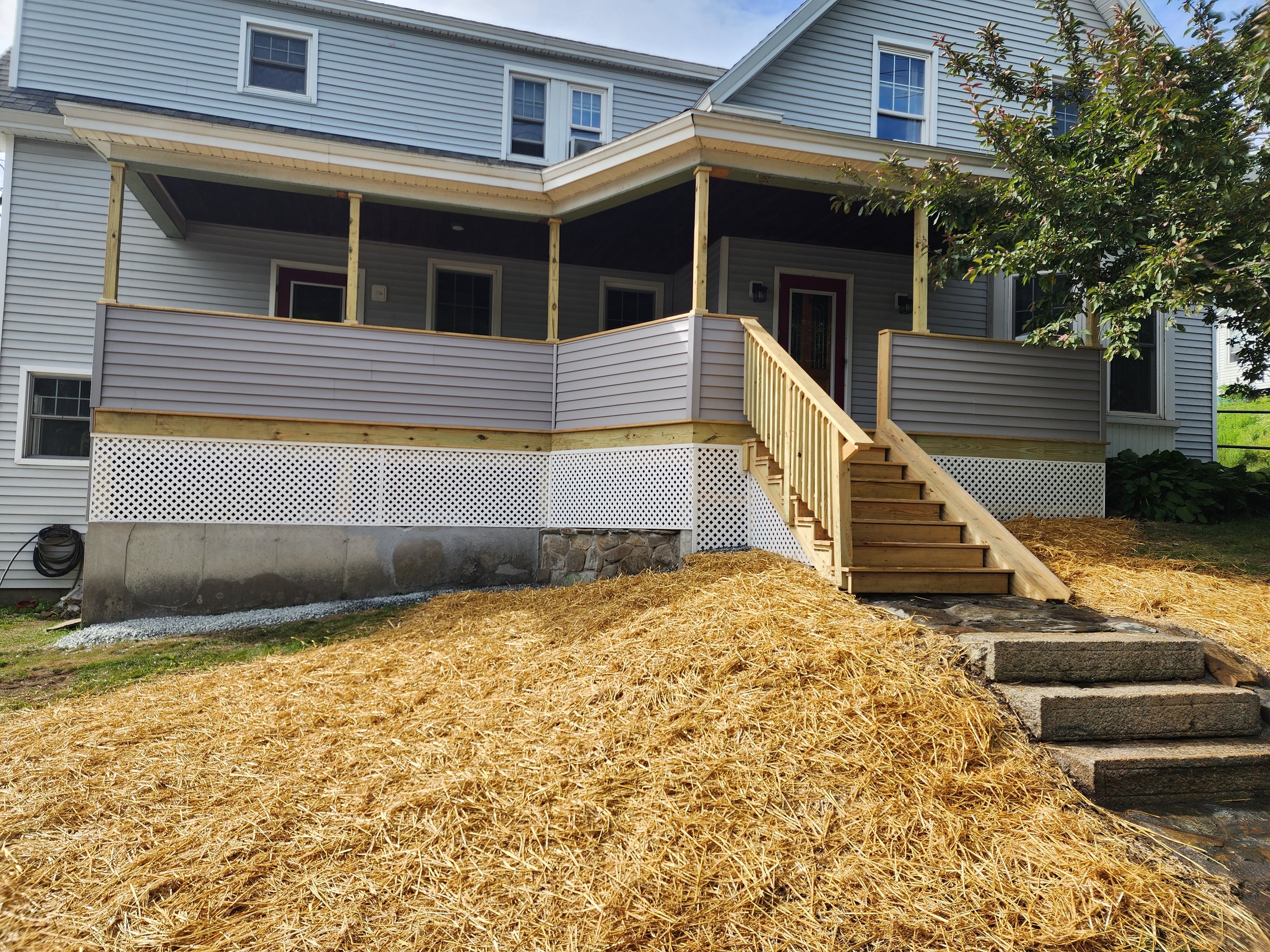 A house with a wooden deck and stairs under construction, with a tree on the right side and a mix of grass and straw on the ground.