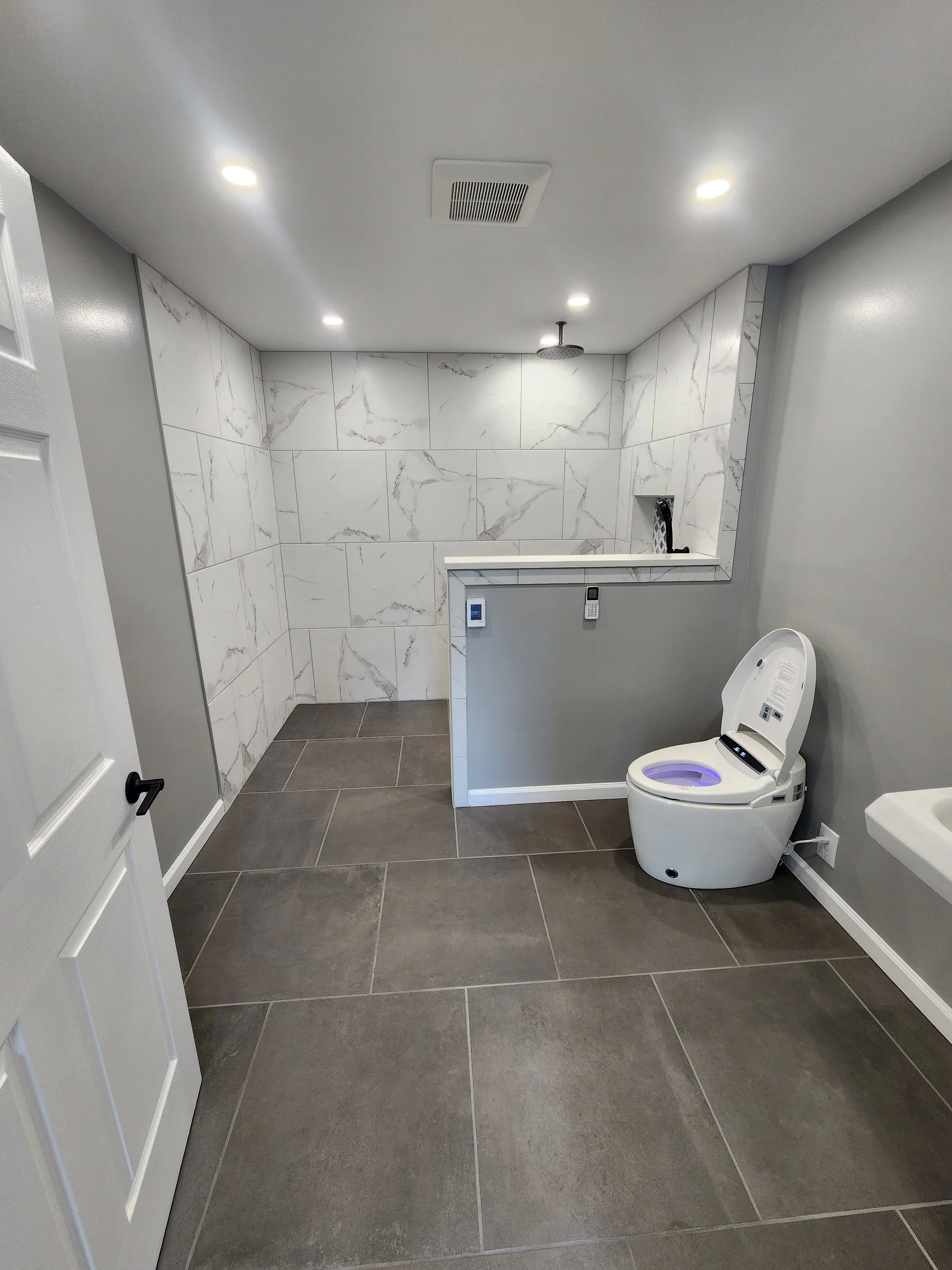 A modern bathroom with white marble tiled walls, gray tiled floor, and gray walls. There is a toilet on the right side with a glowing purple seat, and a small ledge with a black and white checkered cloth in the corner. The ceiling is white with sever