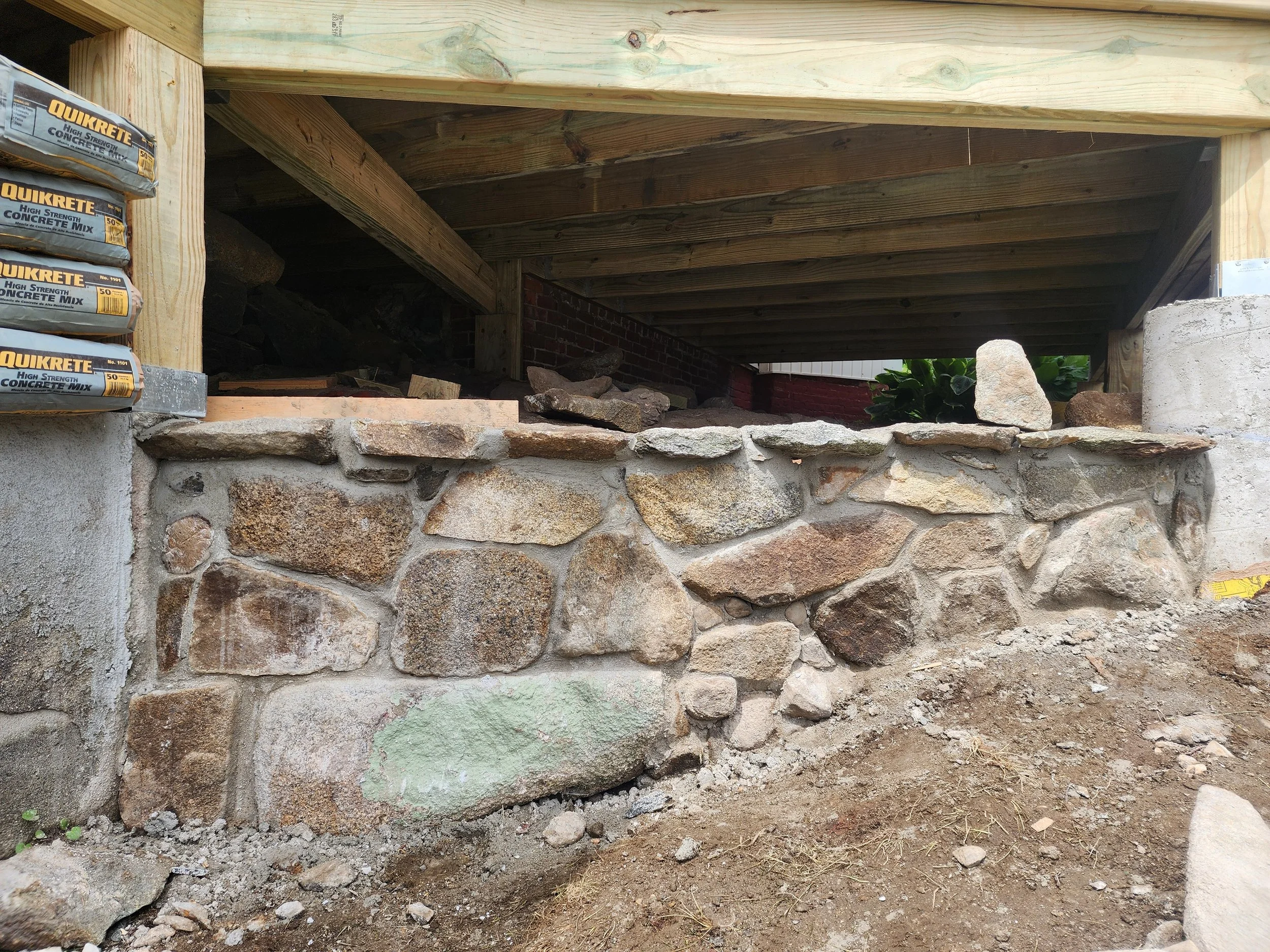 Construction site showing a stone wall base with bricks, a concrete foundation, and wooden framing for a building or deck.