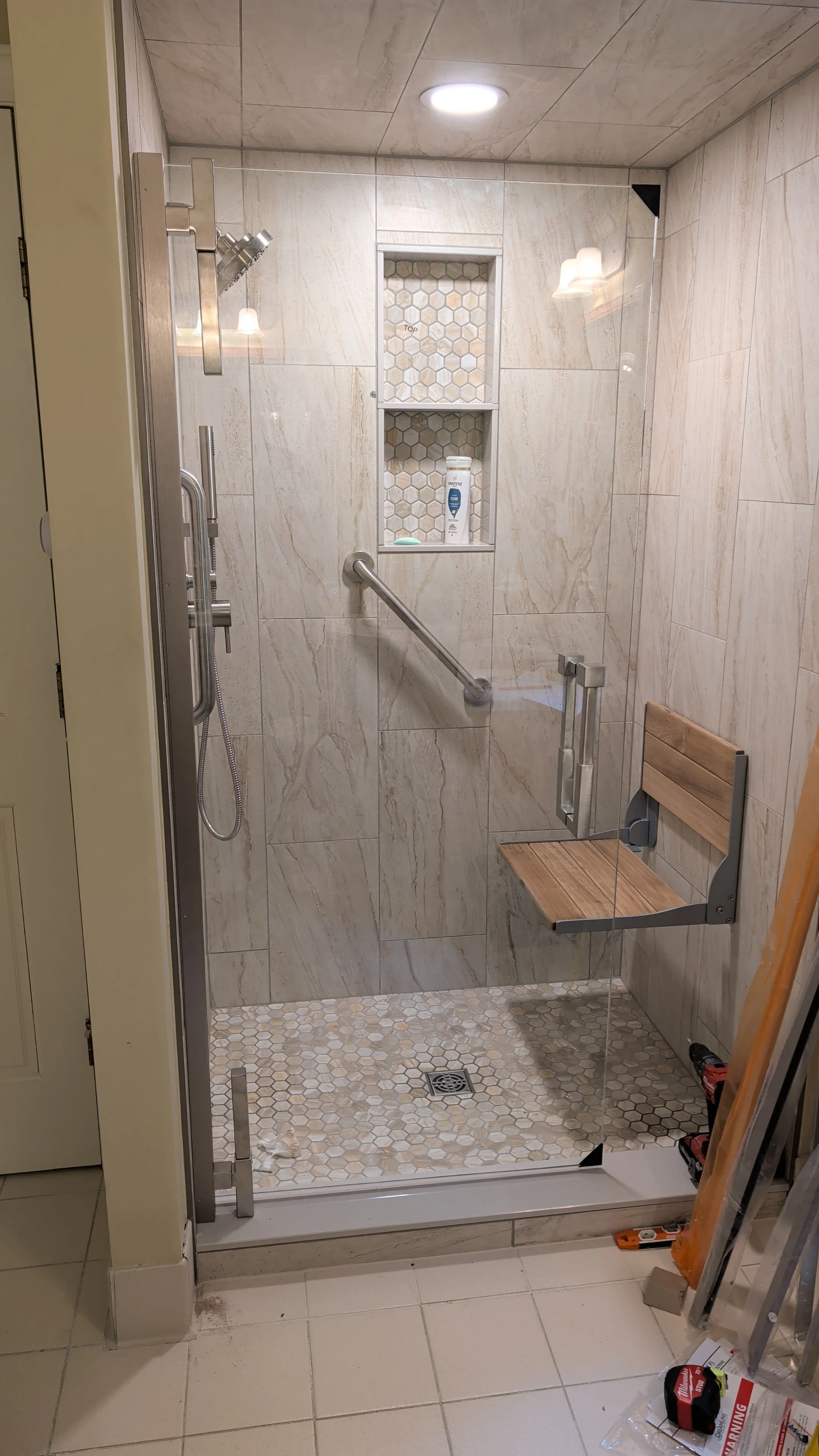 A walk-in shower with beige marble tiles, a built-in shelf with toiletries, a wooden seat, a glass door, and a ceiling light.
