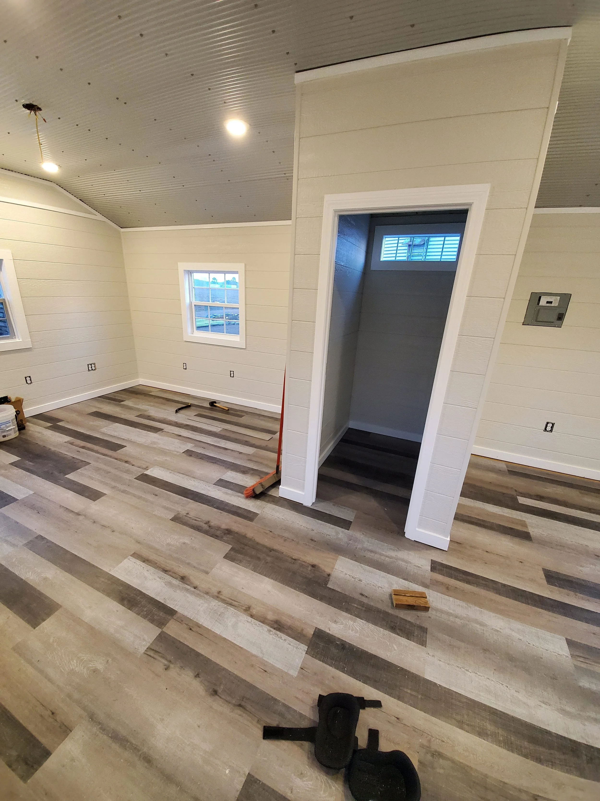 Living room with wood-look flooring, cream-colored horizontal shiplap walls, two small horizontal windows, a partially finished ceiling with two lights and an unfinished ceiling light wiring, and various tools and objects on the floor.