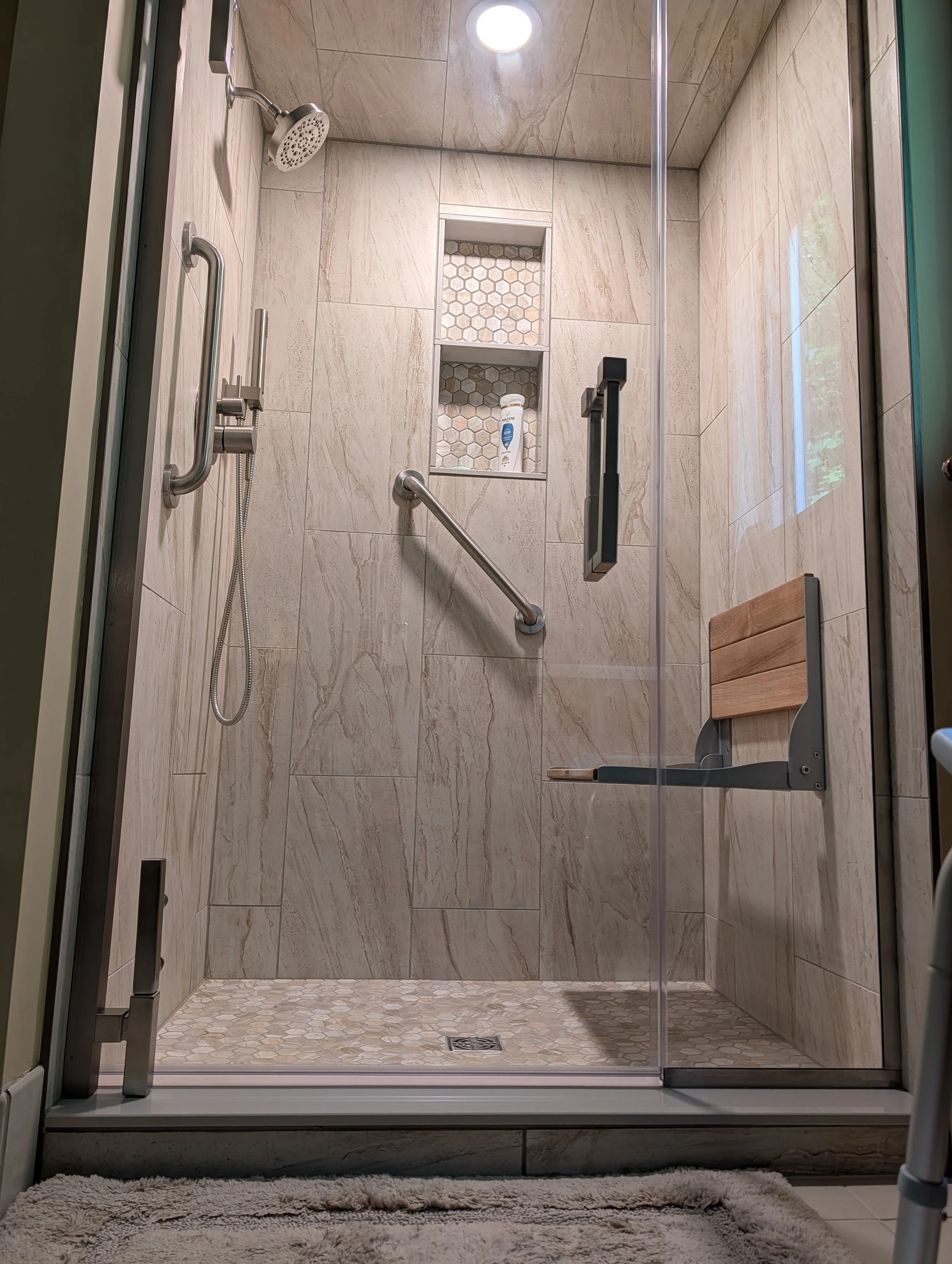 Modern walk-in shower with beige marble tiles, hexagon mosaic tile accents, a built-in niche with toiletries, a rain showerhead, a grab bar, and a wooden shower seat, viewed through a glass door.