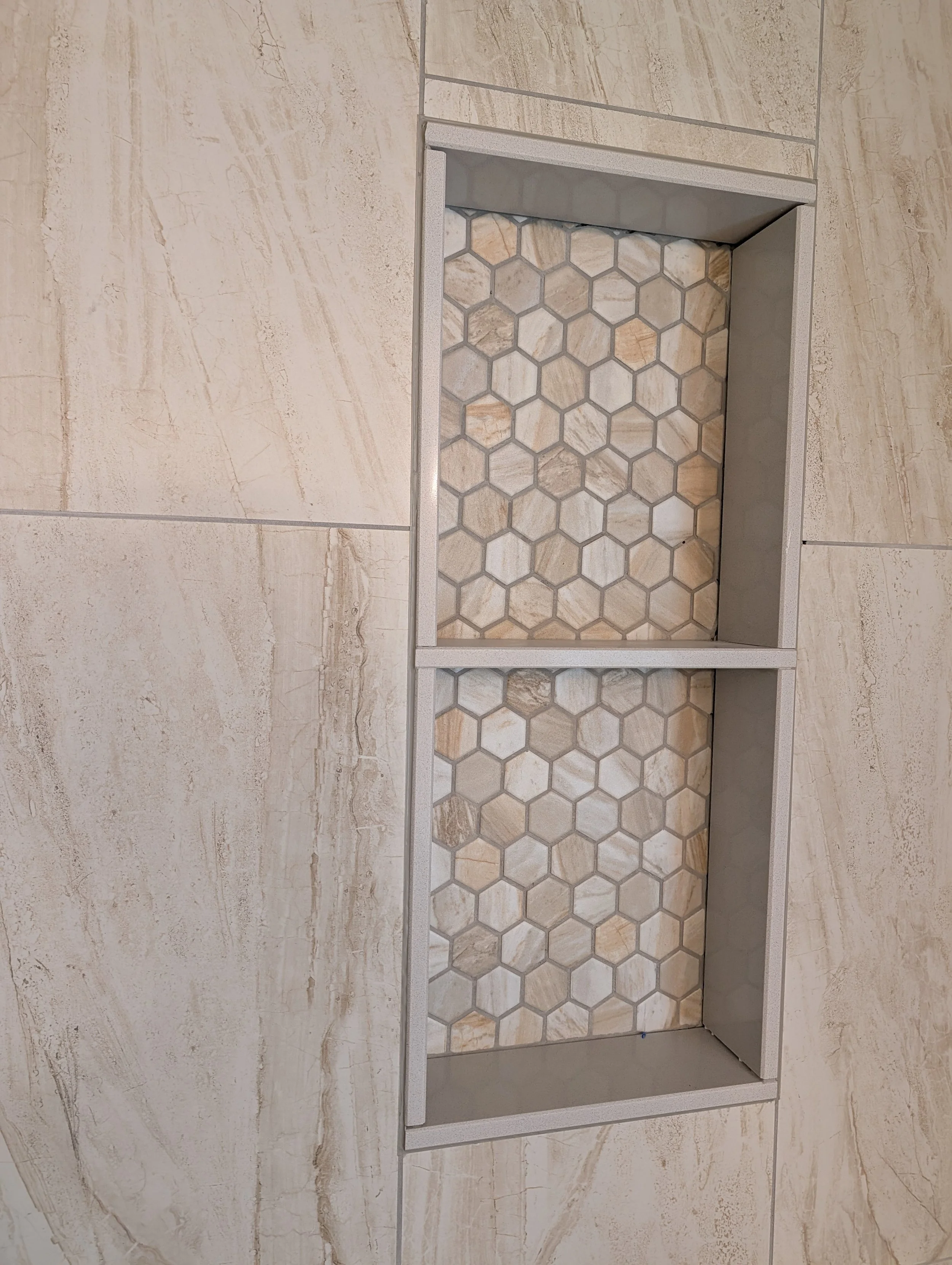 Close-up of a recessed shelf with hexagonal marble tiles inside a bathroom or kitchen wall, bordered by light gray grout and surrounded by beige marble tiles.