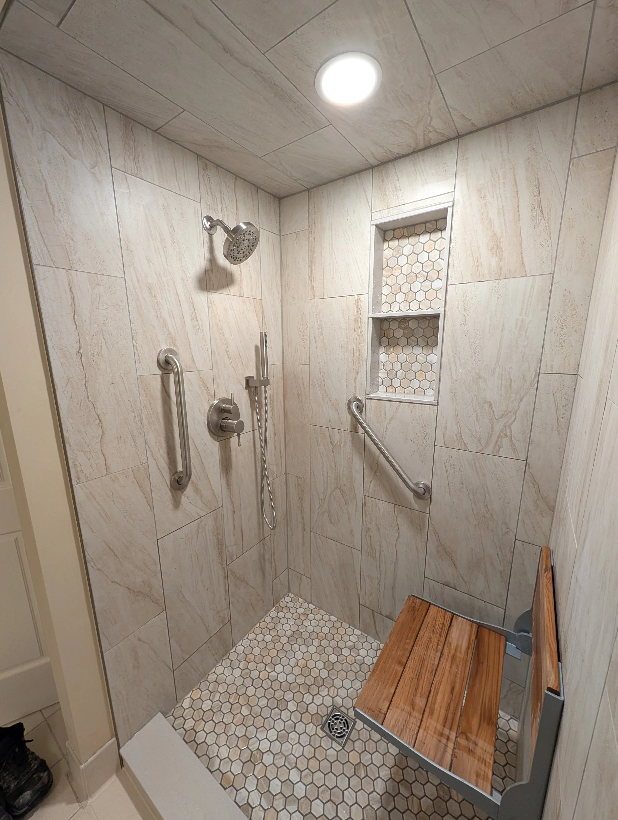 A walk-in shower with beige marble tiles on the walls and hexagon tile on the floor. It has a showerhead, a handheld shower attachment, and a grab bar. There are two built-in niches with hexagon mosaic tile. A wooden bench is positioned in the shower