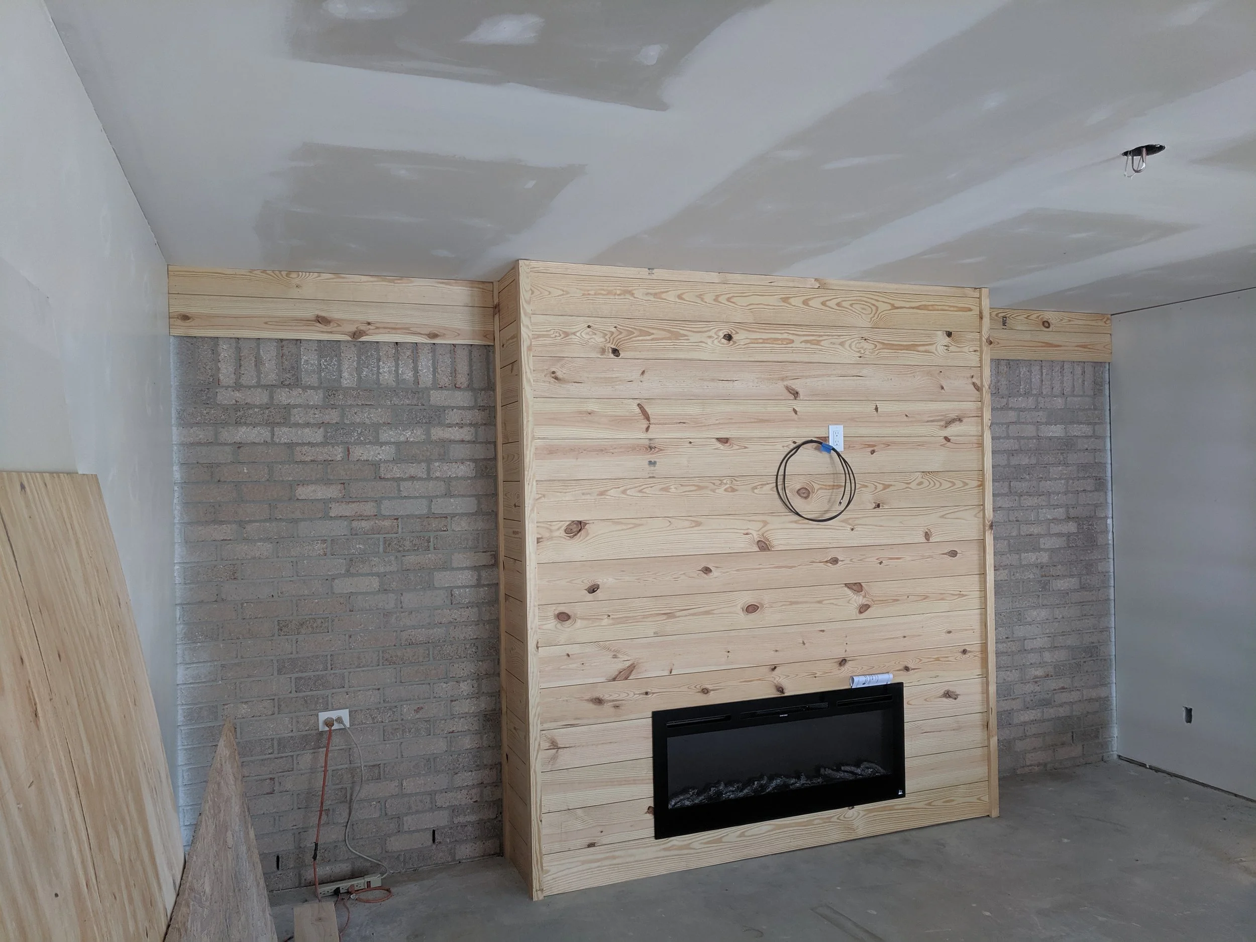 Interior of a room under construction, featuring a wooden frame wall with a fireplace, exposed brick wall, electrical outlets, and wiring.