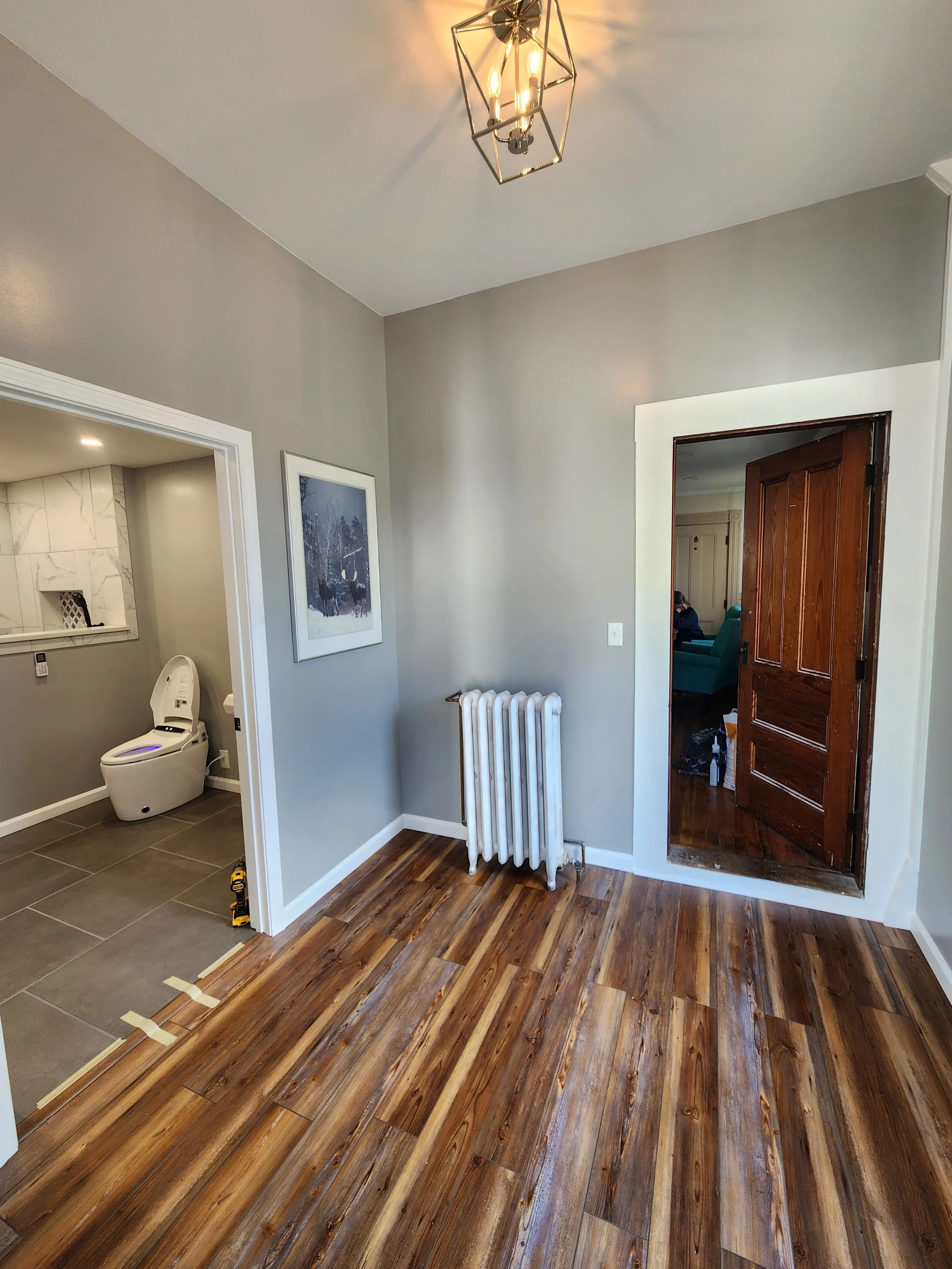 Interior of a house showing a hardwood floor, a radiator, a doorway to another room, and a nearby bathroom with a modern toilet. The walls are light gray, and there is a framed picture hanging on the wall. A ceiling light fixture is visible.