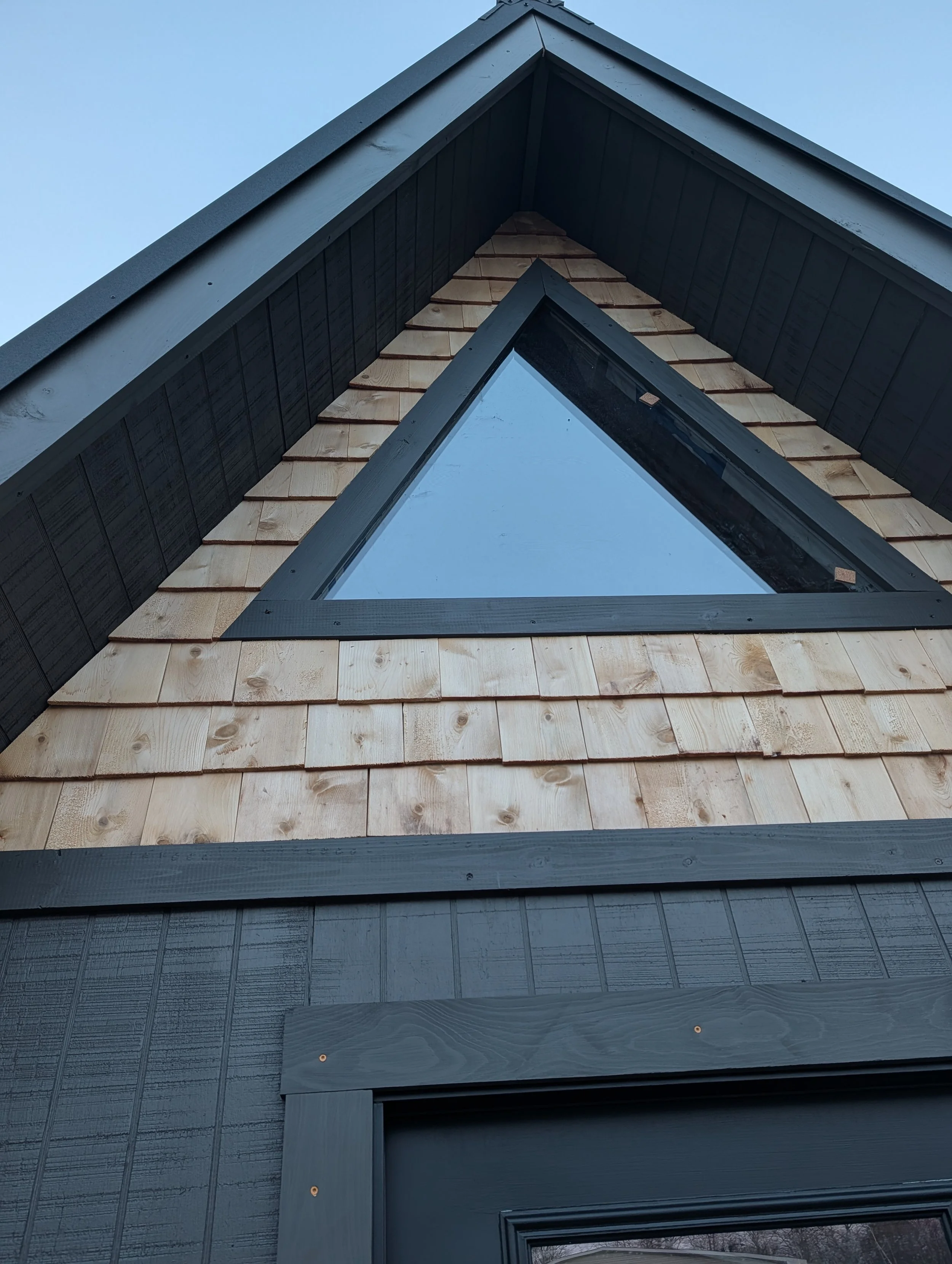 Close-up of a house roof with black and natural wood-shingled sections, with a triangular skylight window in the center, and a light blue sky in the background.