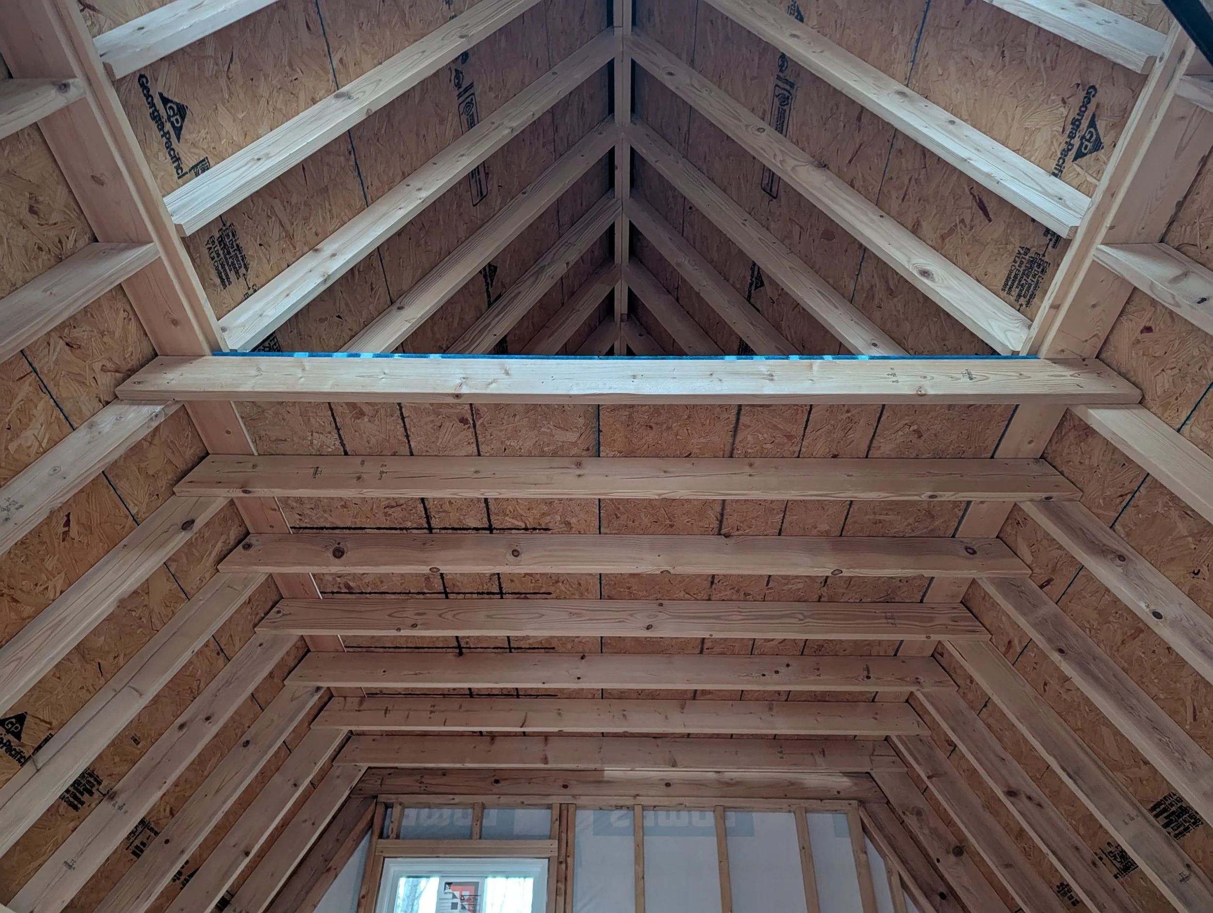The photo shows the interior of a building under construction with exposed wooden trusses and plywood sheathing on the walls. There is an opening at the far end showing a glimpse of the outside.