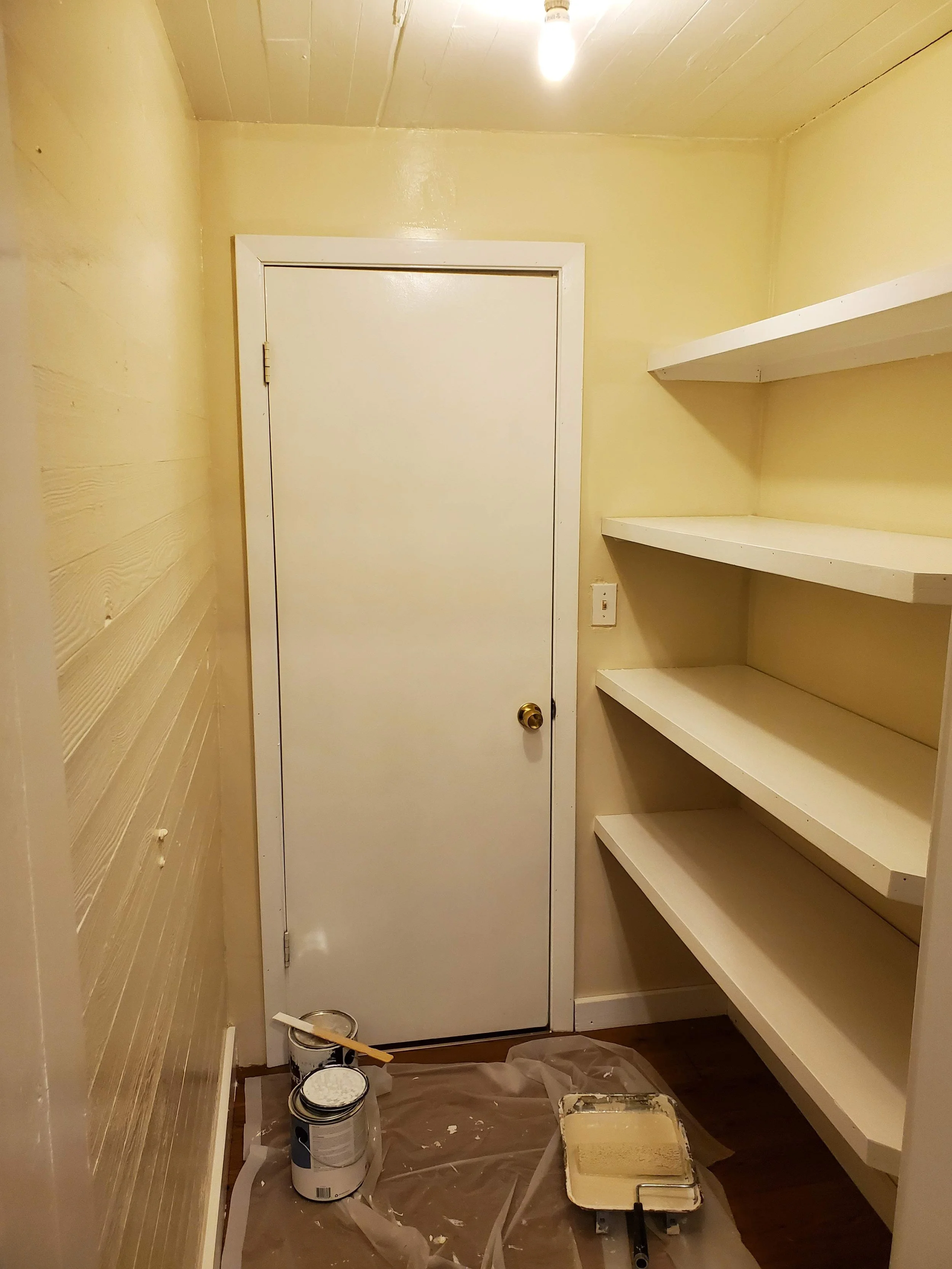 A small room being painted with beige color. There are open paint cans, painting tools, and plastic sheets on the floor. Shelves are installed on the right wall, and a closed door with a brass doorknob is on the left wall.