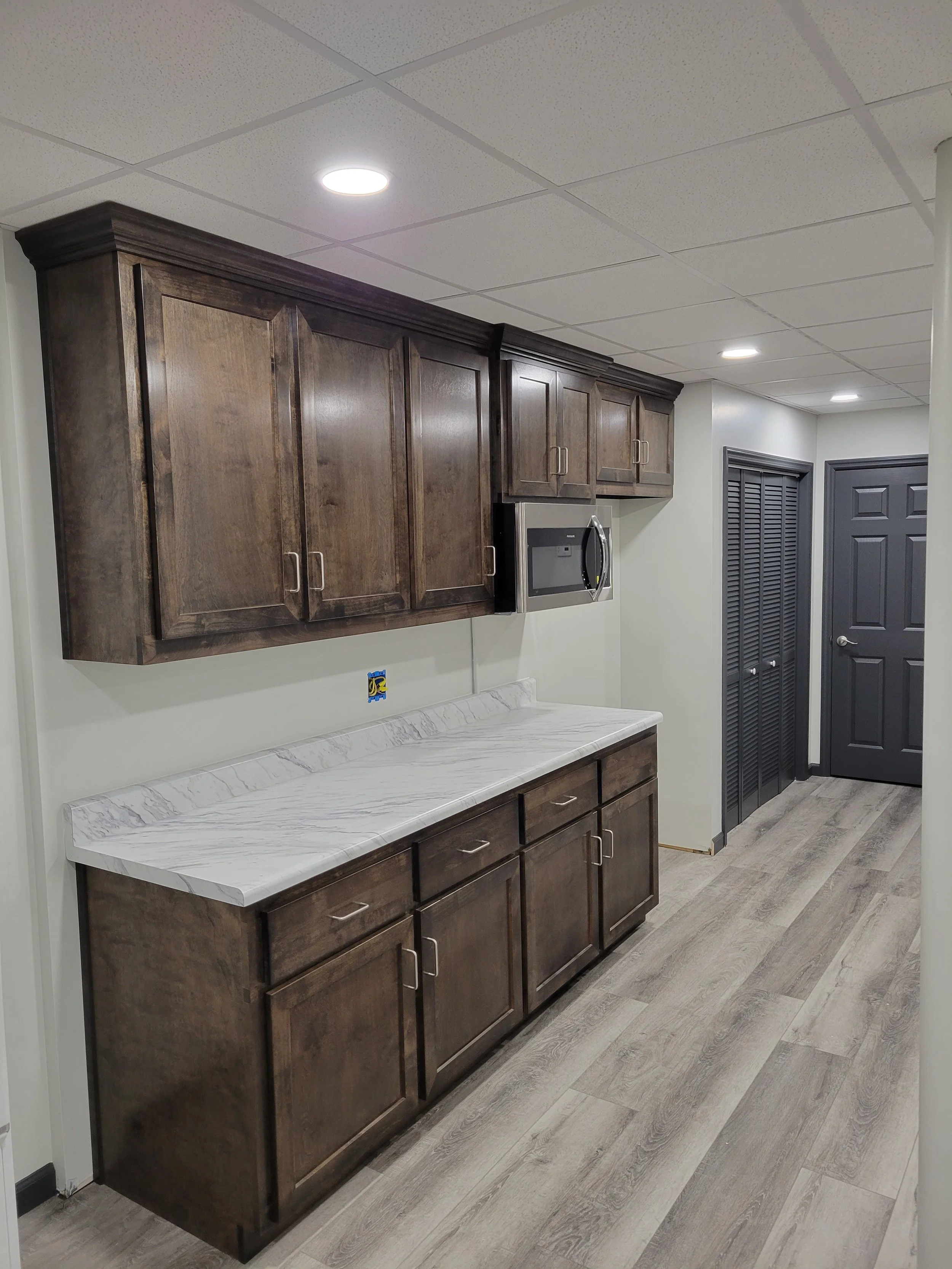 Kitchen with dark wooden cabinets, white marble countertop, microwave oven, gray door with louvered panels, and wood-look flooring.