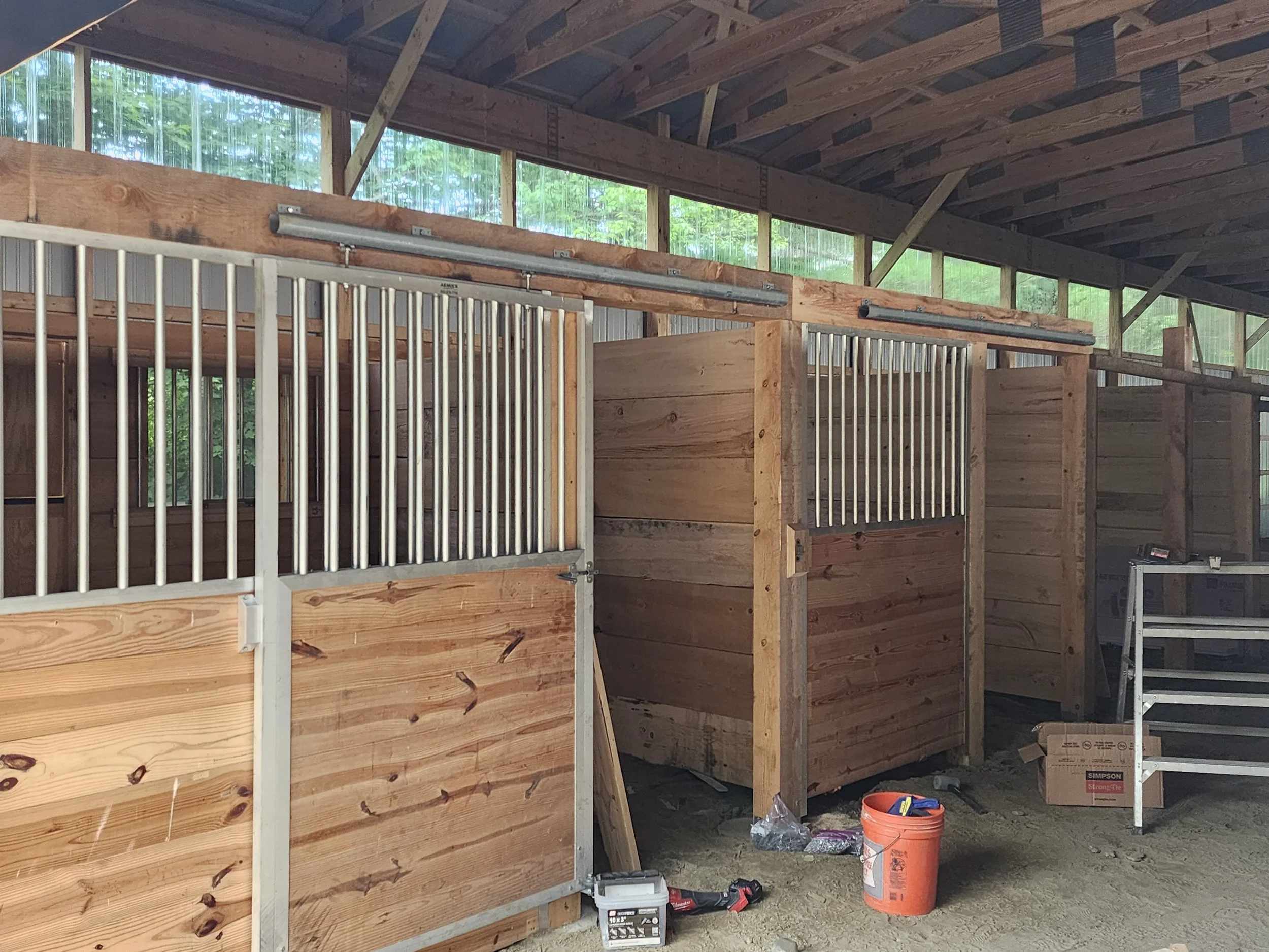 Construction site with wooden partitions and metal framing inside a building under construction. Construction tools and equipment pile on the unfinished floor, natural light filtering through window openings.