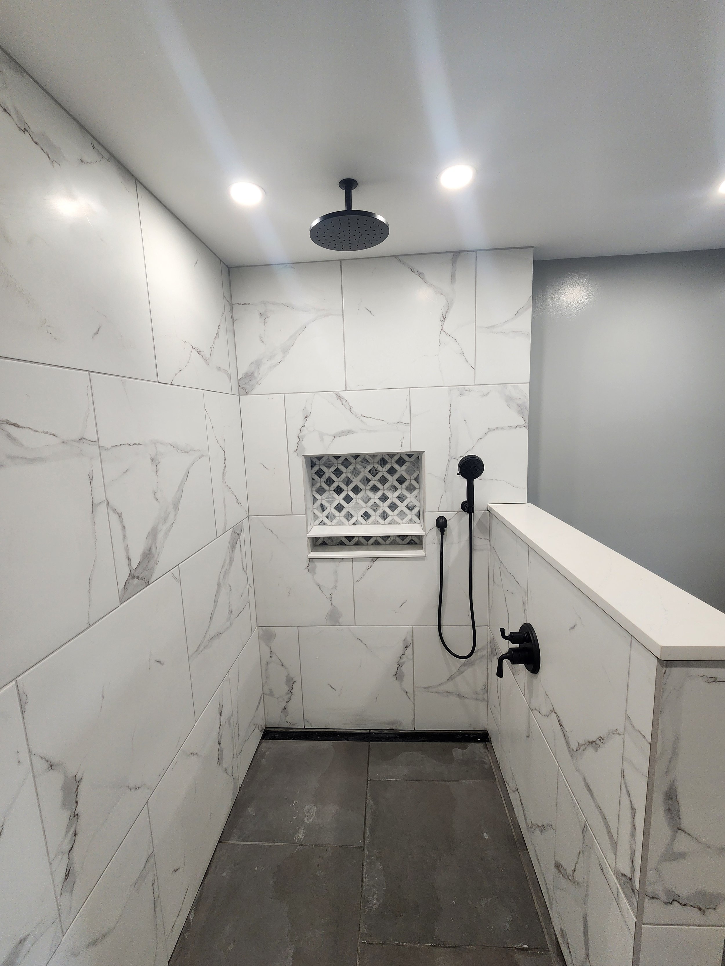 A modern walk-in shower with white marble tiles on the walls and dark gray tiles on the floor. There is a round rain showerhead mounted on the ceiling, a handheld shower spray, and a built-in niche with decorative tiles.