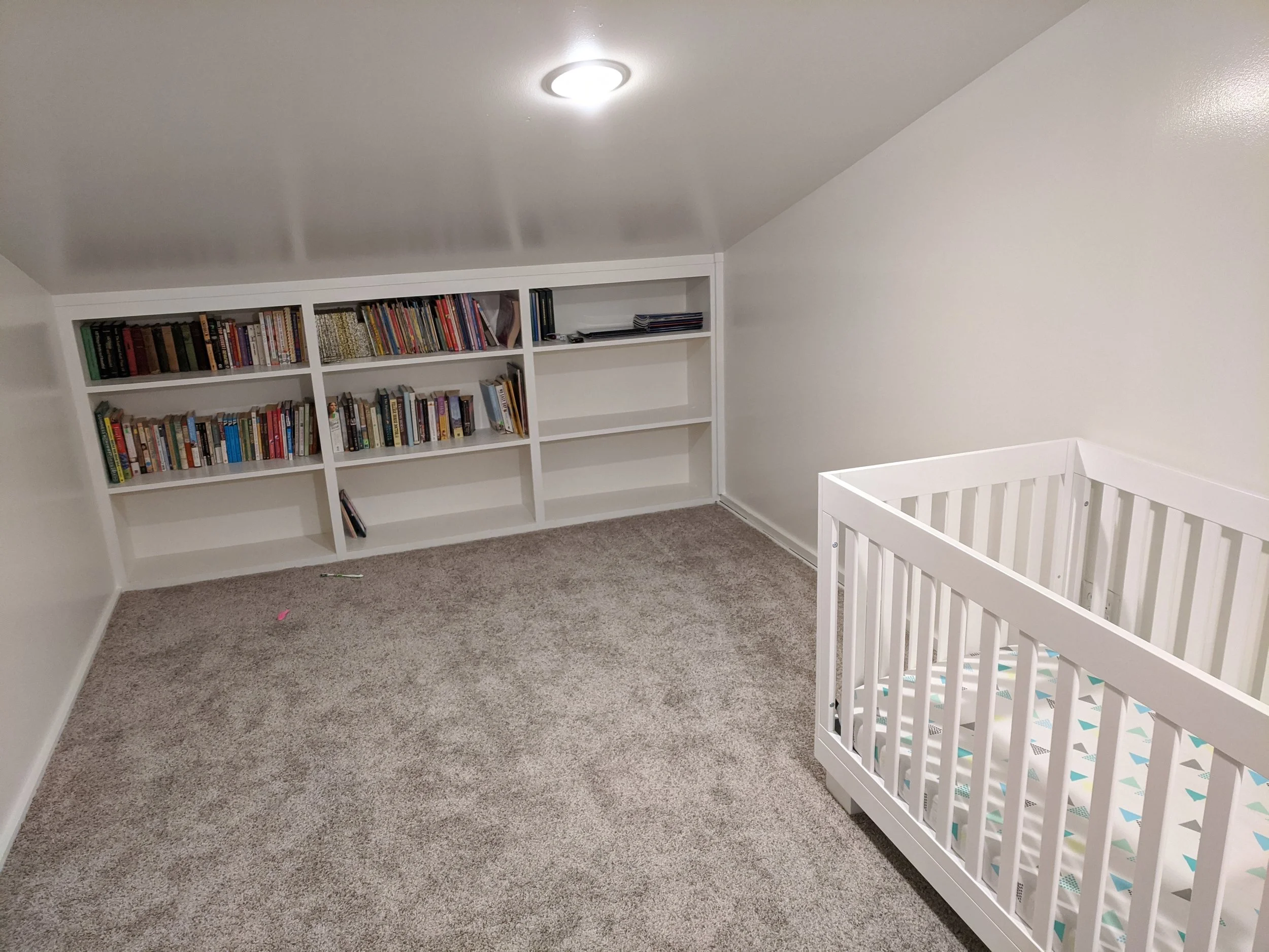 A corner of a room with white bookshelves filled with books and magazines, a white baby crib, and beige carpeted flooring.