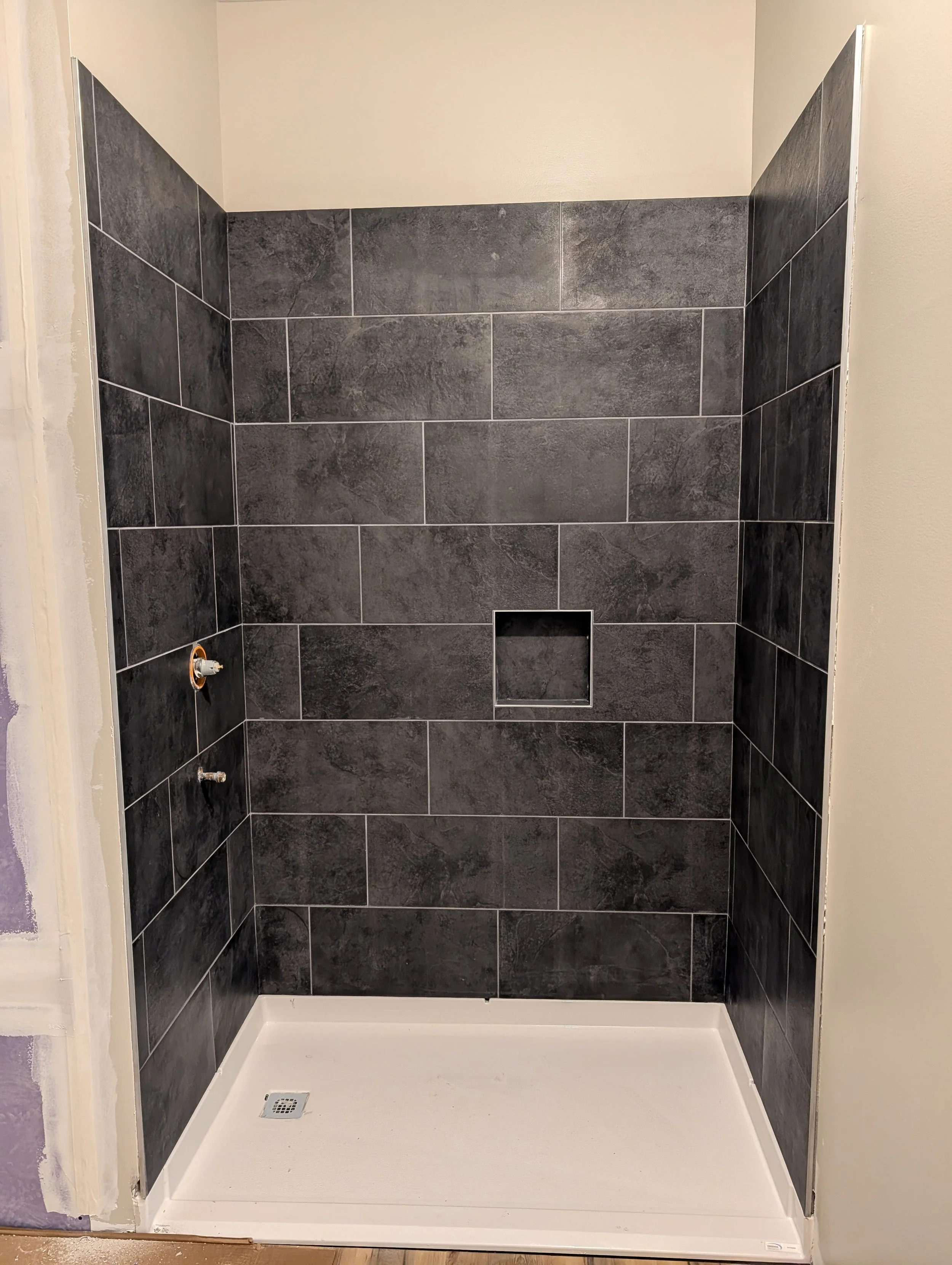 Bathroom shower with black tile walls, a white shower base, a drain, and an unfinished wall with visible drywall on the side.