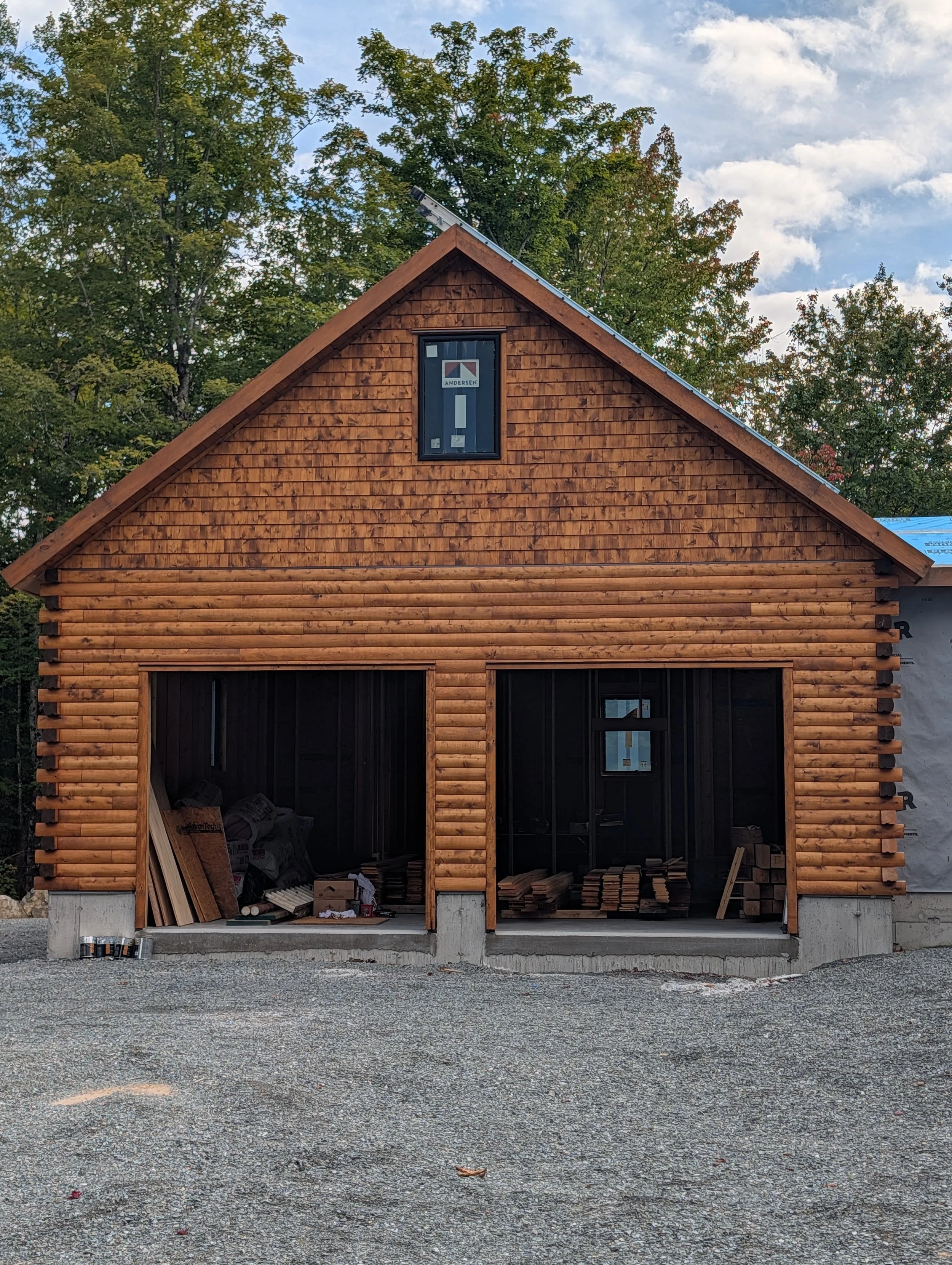 A house under construction with wooden siding, two open garage doors, construction materials inside, and a small window near the roof with the brand name Andersen. The house has a concrete foundation and is surrounded by trees with green leaves, unde