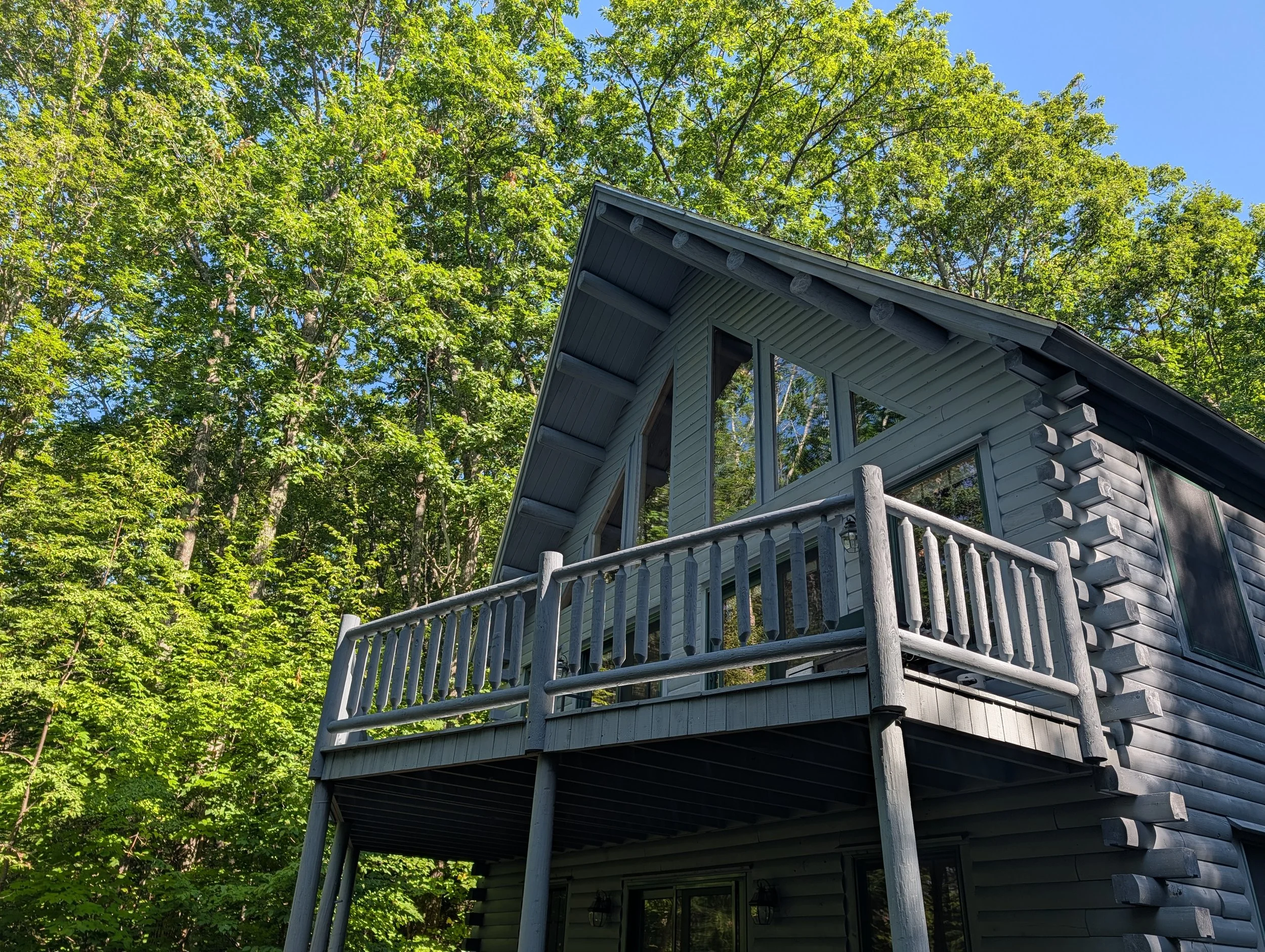 A house with gray siding and large windows has a wooden deck with a railing. It is surrounded by tall green trees and a bright blue sky.