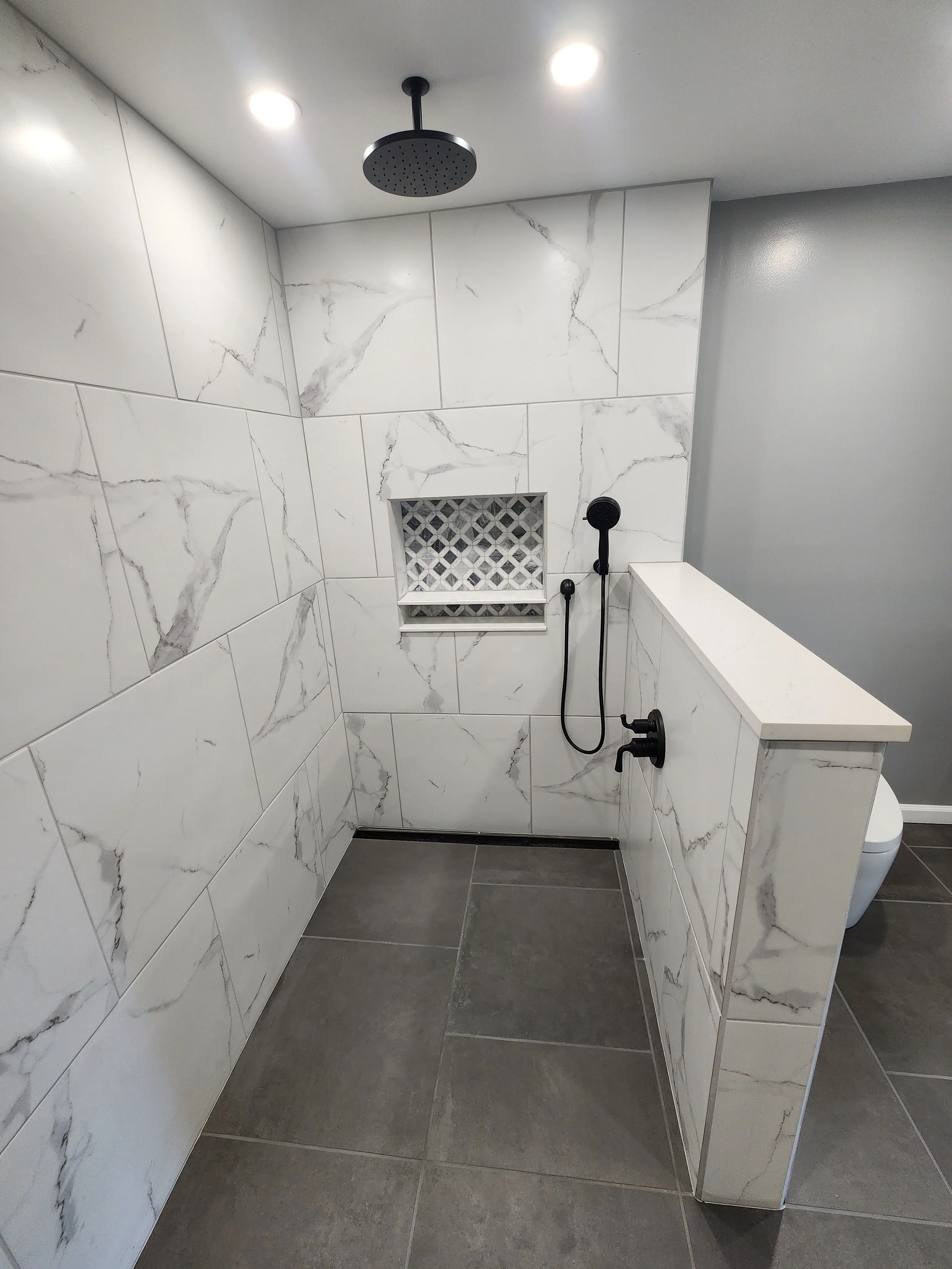 A modern walk-in shower with white marble tiles and gray grout. It features a round rainfall showerhead on the ceiling, black handheld shower, and a built-in niche with a geometric tile pattern. There is a half-wall with marble tiling dividing the sh