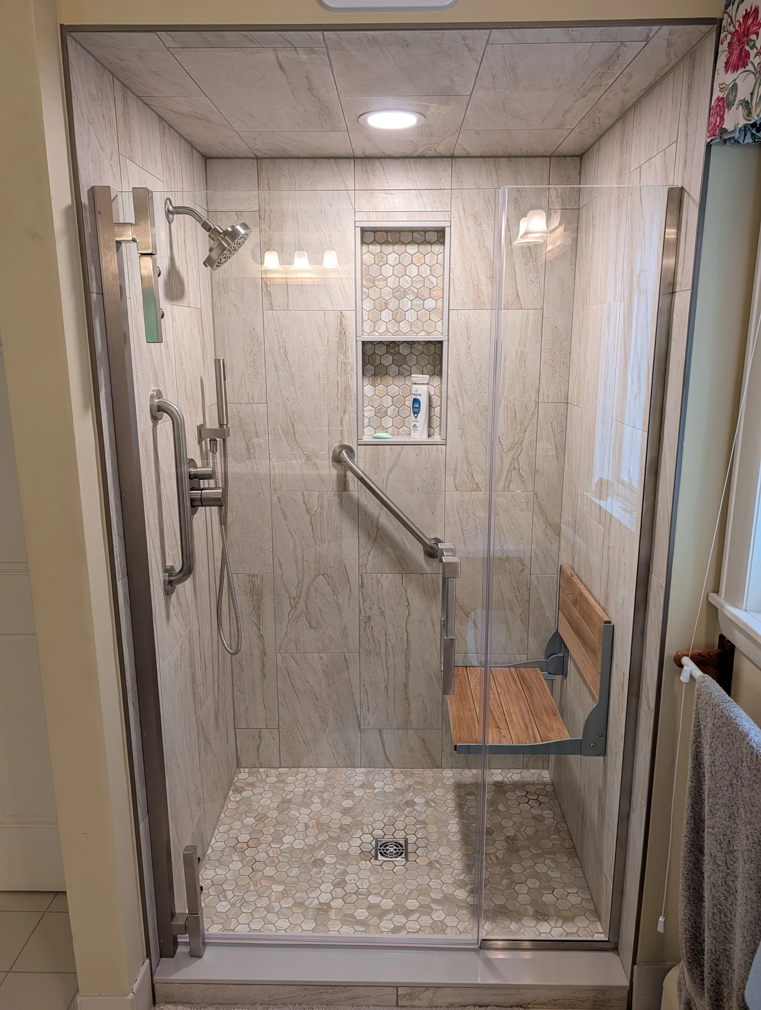 A modern walk-in shower with beige marble tiles, hexagonal floor tiles, a built-in niche for toiletries, a ceiling light, and a glass door with a metal handle.