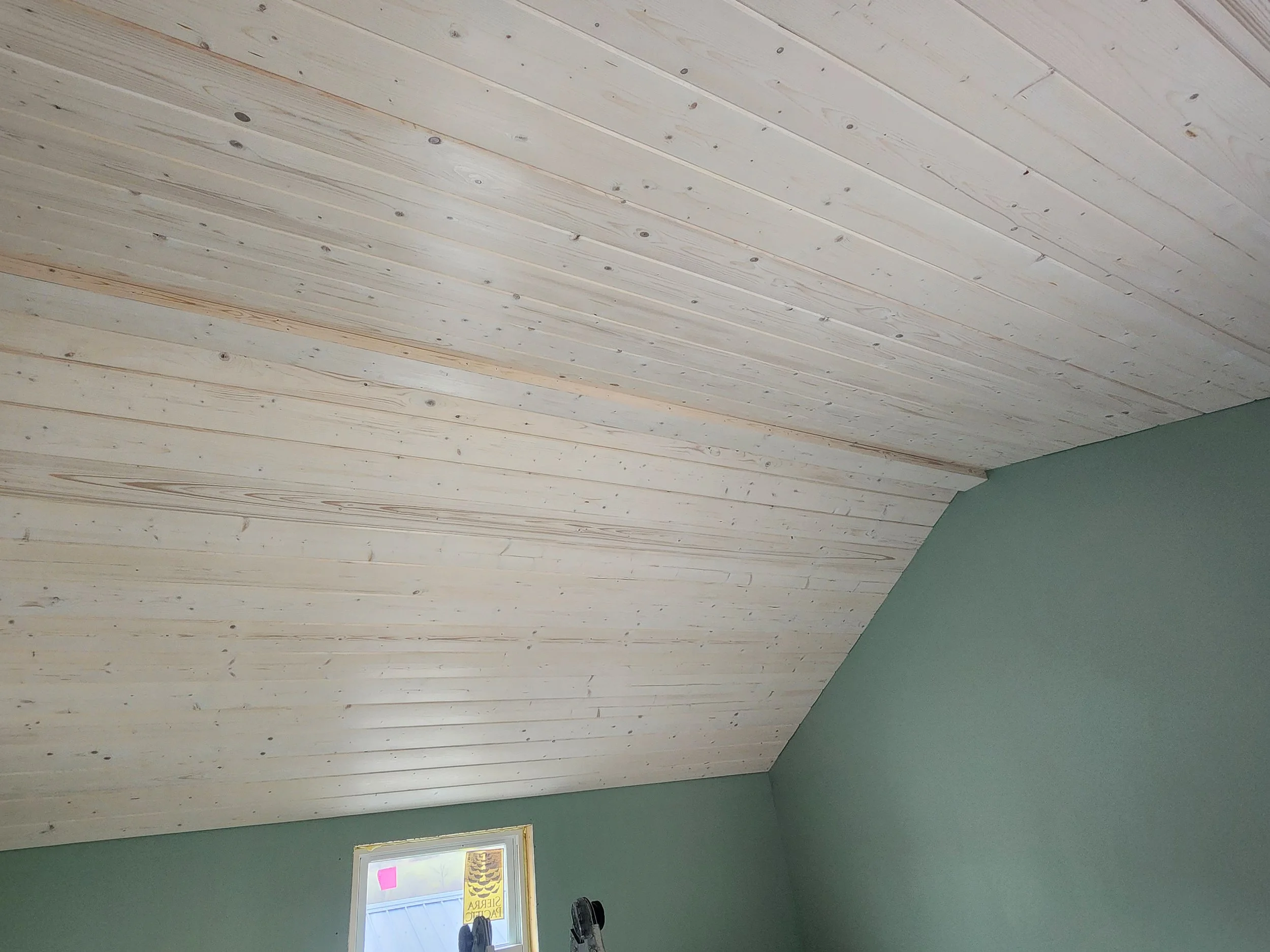 Interior view of a room with a wooden plank ceiling and green walls, a small window with a notice on it, and some black objects near the window.