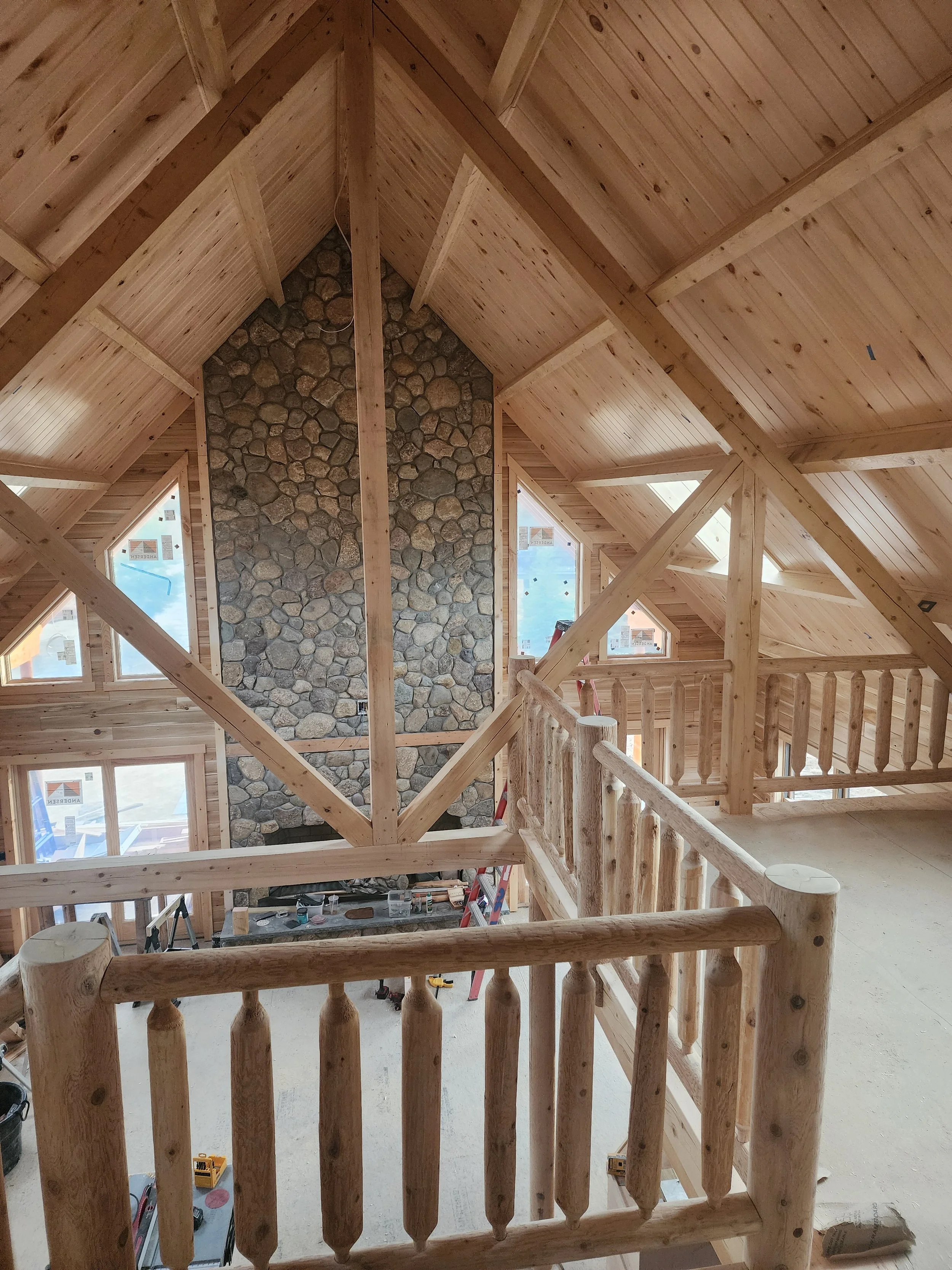 Interior view of a wooden loft under construction with a stone fireplace, wooden railings, and multiple windows providing natural light.