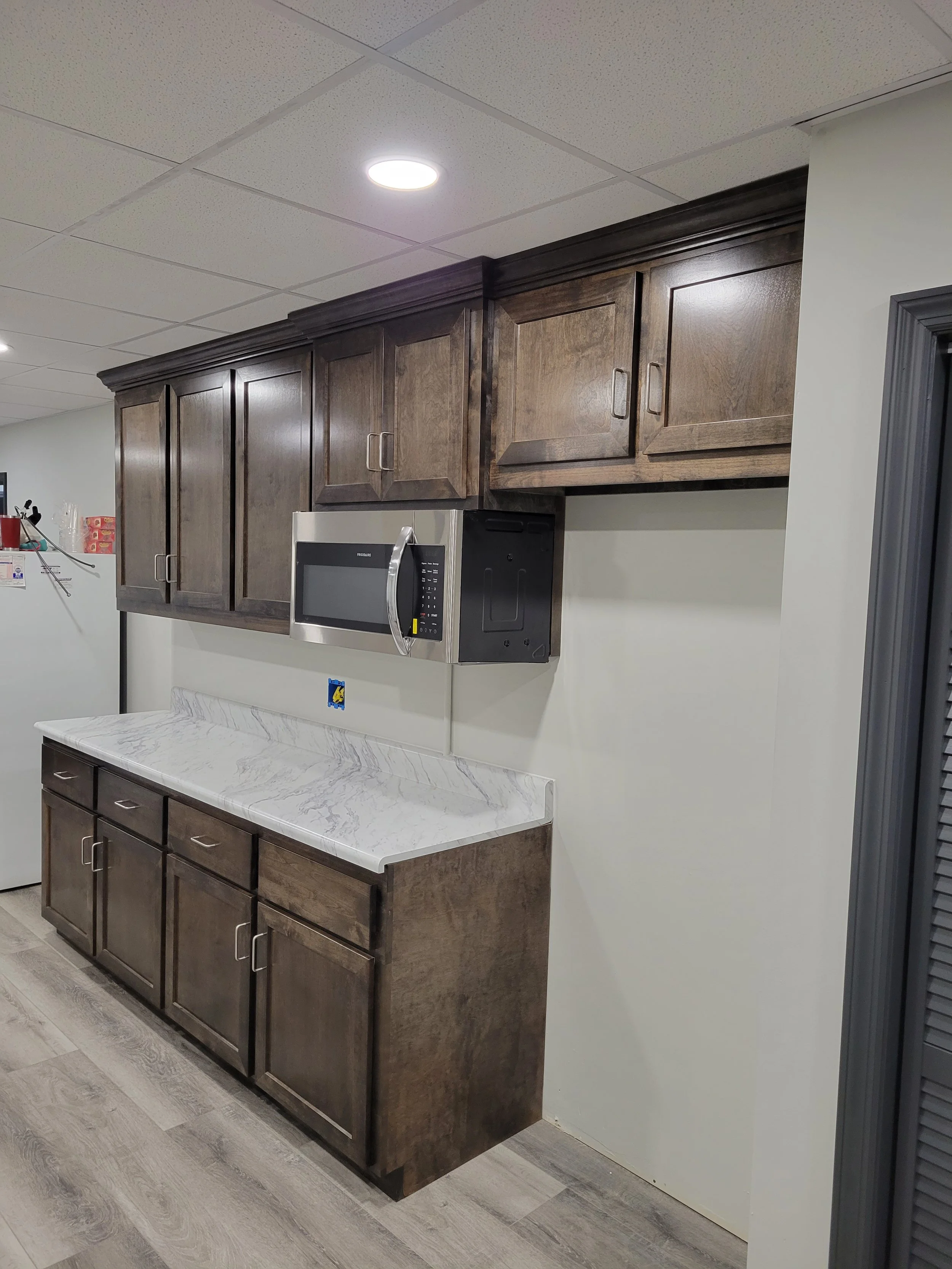 Kitchen area with dark wooden cabinets, a microwave oven, a marble countertop, beige walls, and gray wood flooring.