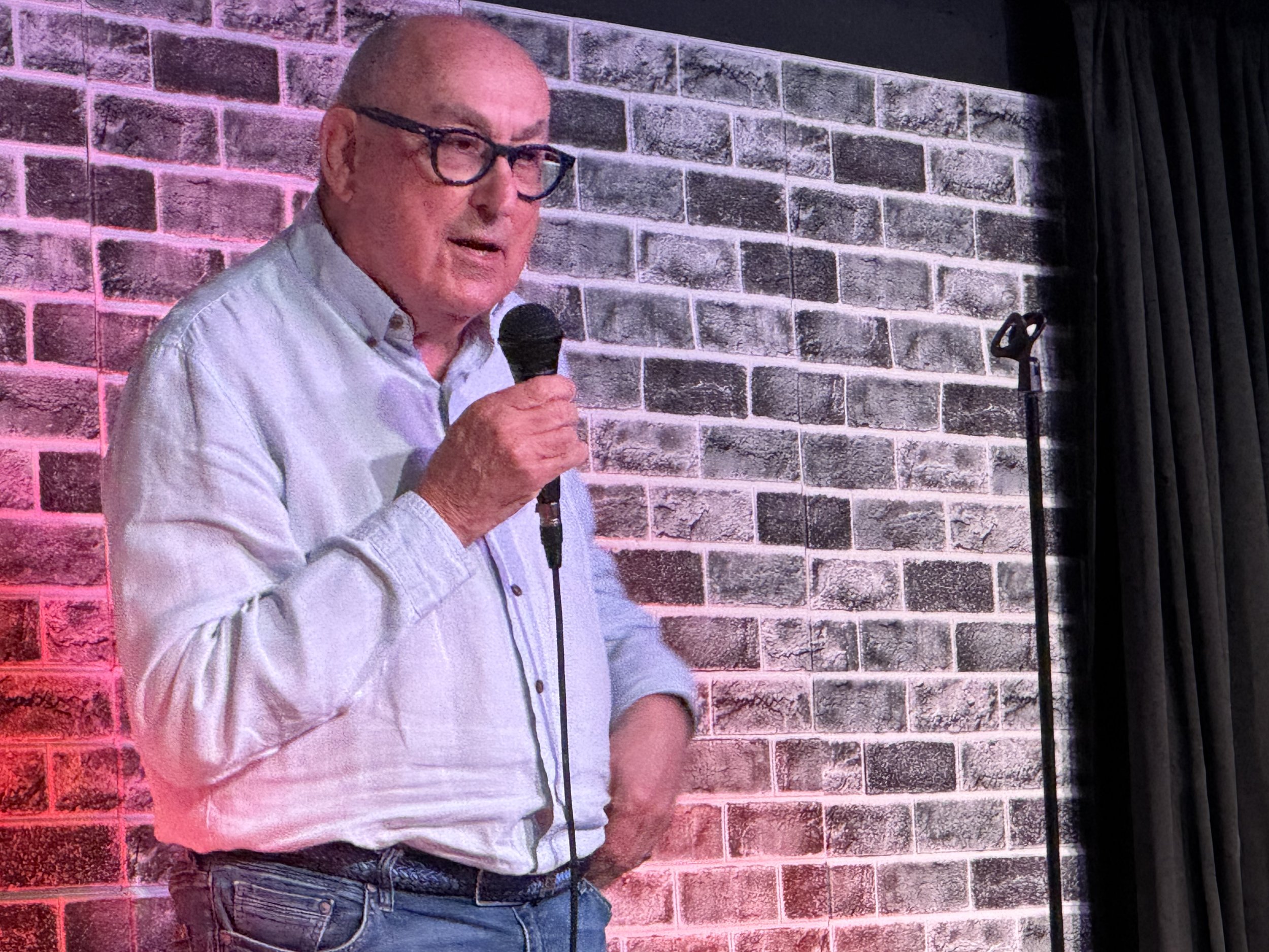 A man with glasses, wearing a light-colored shirt and jeans, is holding a microphone and speaking, standing against a brick wall background.