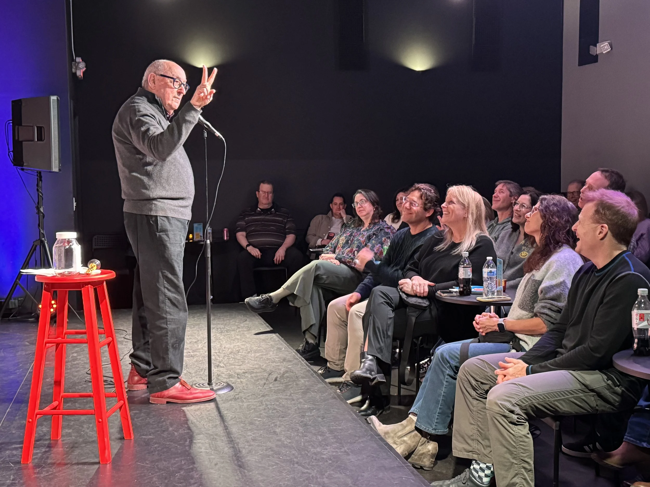 An elderly man with glasses and a grey sweater giving a speech on stage to an audience. The man is making a peace sign with his right hand. The stage has a red stool and a microphone. The audience is seated and appears engaged, with some smiling and others listening attentively.
