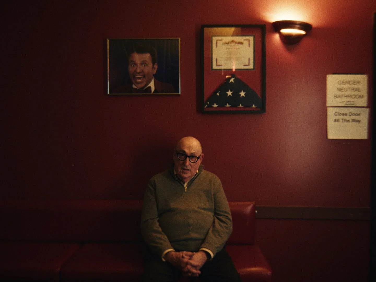 An elderly man with glasses sitting on a red cushioned bench against a dark red wall, with framed pictures and a wall lamp behind him.