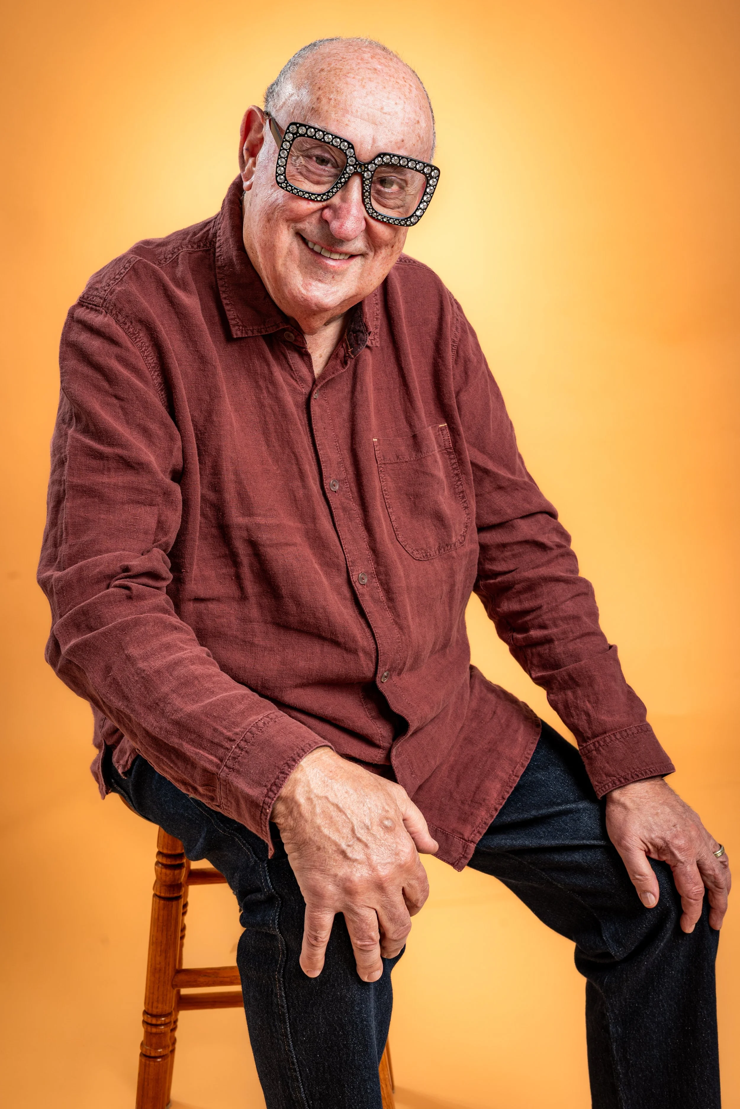 An older man with a bald head and freckles wearing large black glasses with rhinestones, a maroon button-up shirt, and dark jeans, sitting on a wooden stool against an orange background.
