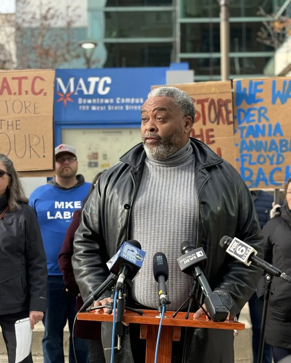 Man speaking at a podium with multiple microphones during a public event, surrounded by people and protest signs about social justice and police accountability.