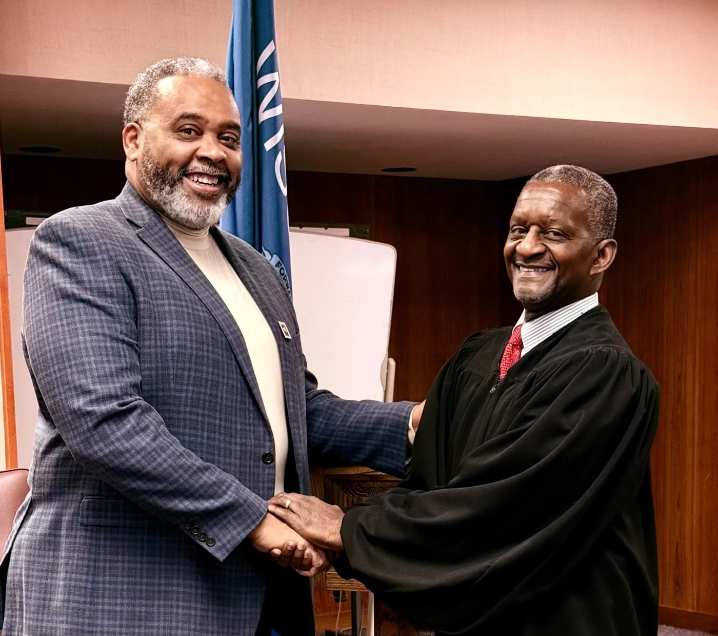 Two men shaking hands, one in a suit and the other in a graduation gown, smiling in an indoor setting with a flag in the background.