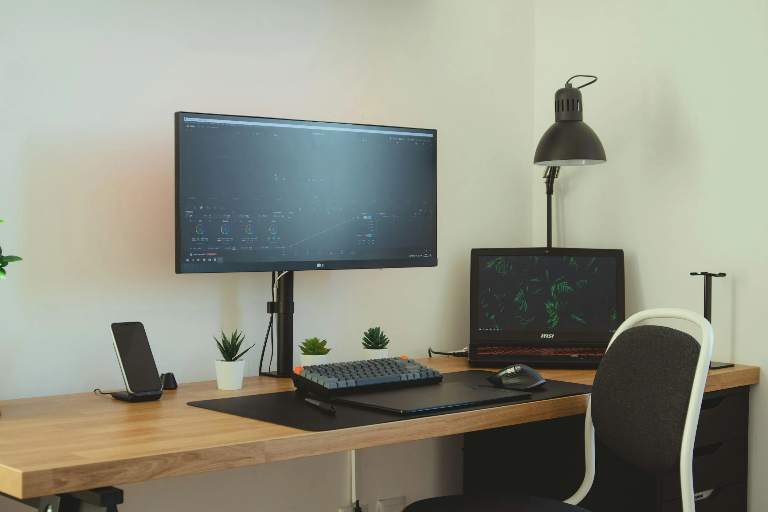 A home office setup with a wooden desk, a large computer monitor with editing software, a smaller laptop displaying a tropical leaf pattern, a gaming keyboard, a mouse on a mouse pad, a smartphone on a stand, and three potted succulents. There is a black adjustable desk lamp and a white and grey office chair. The background features a plain light-colored wall.