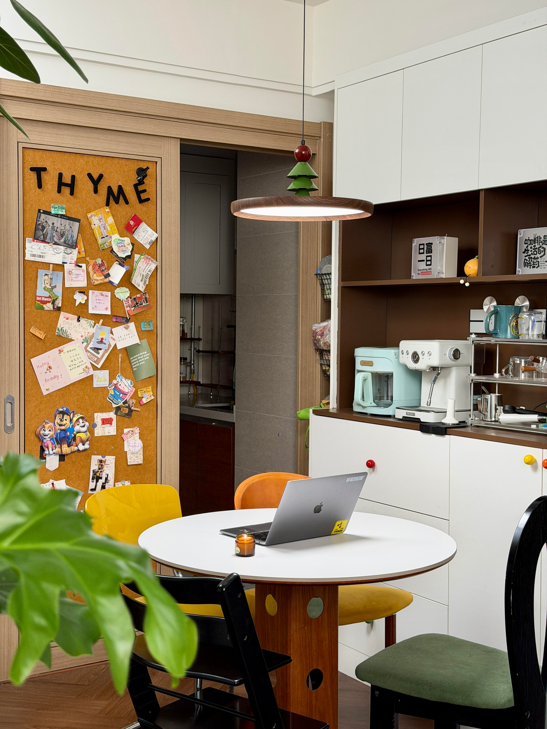 A modern kitchen and dining area with a white round table, black and yellow chairs, a laptop, a small candle, a coffee machine, shelves with books, cups, and kitchen items, and a bulletin board with notes and decorations. A green plant partially visible in the foreground.