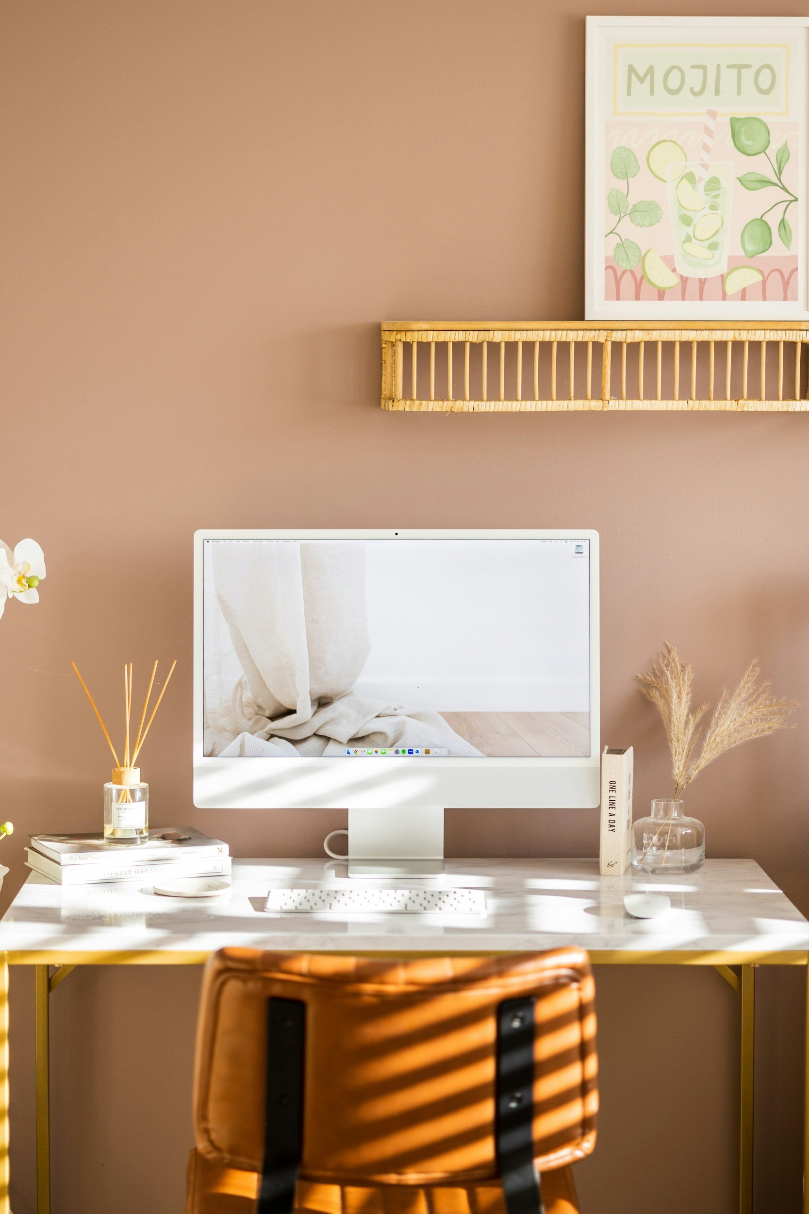 A white computer monitor on a desk with a beige wall background. The screen displays a neutral, crumpled fabric. To the left of the monitor, there is a glass vase with a white flower and dried grasses, and a small stack of books. To the right, there is a closed book, a clear vase with dried grasses, and a small decorative object. A chair with a wooden backrest is in the foreground, partially visible.